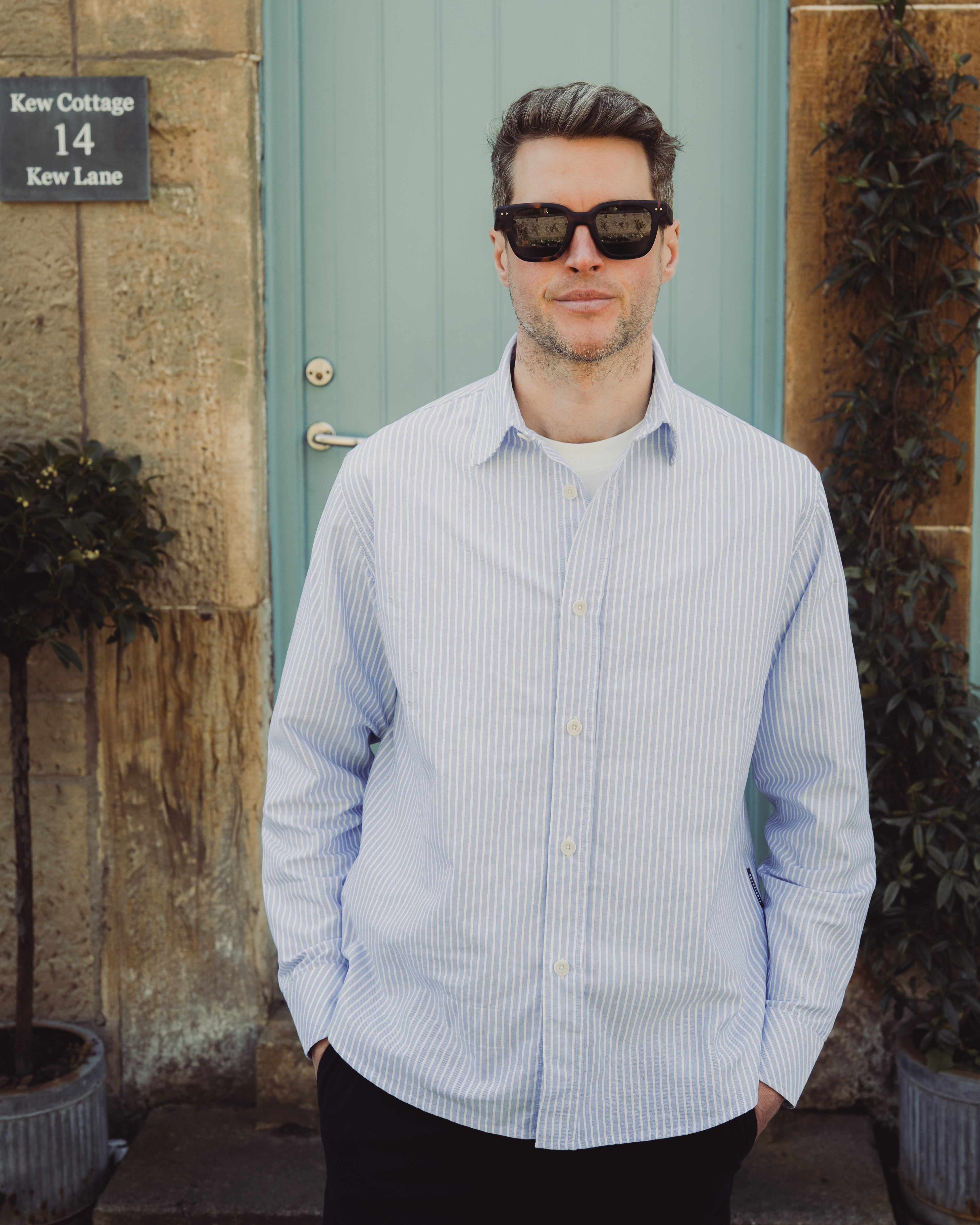 Man wearing sunglasses and a light blue striped shirt standing in front of a building with a door and plants.
