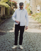 Man in light blue shirt and black pants standing on a cobblestone path with greenery around.