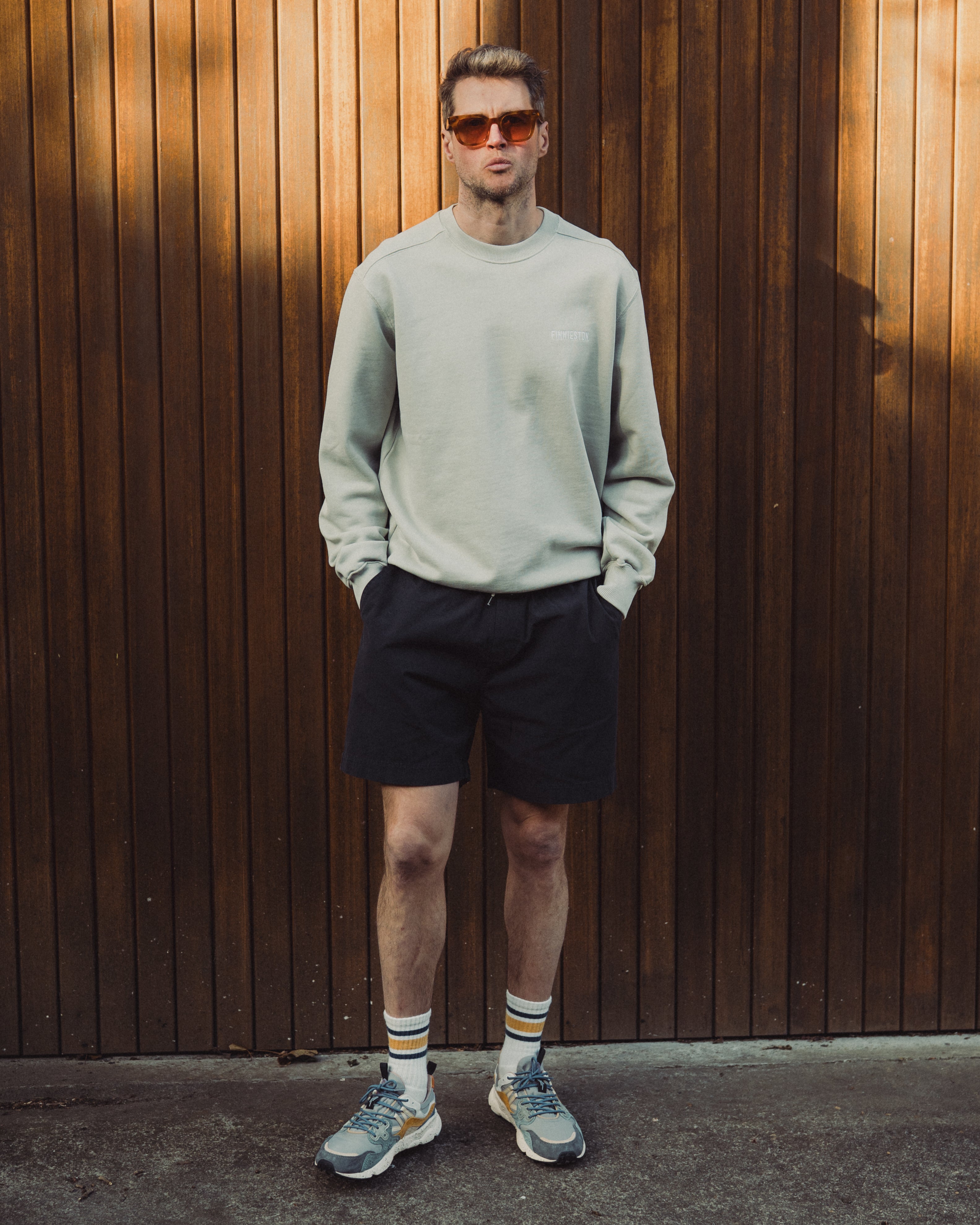 Man wearing a light gray sweatshirt, black shorts, and sunglasses standing against a wooden wall.