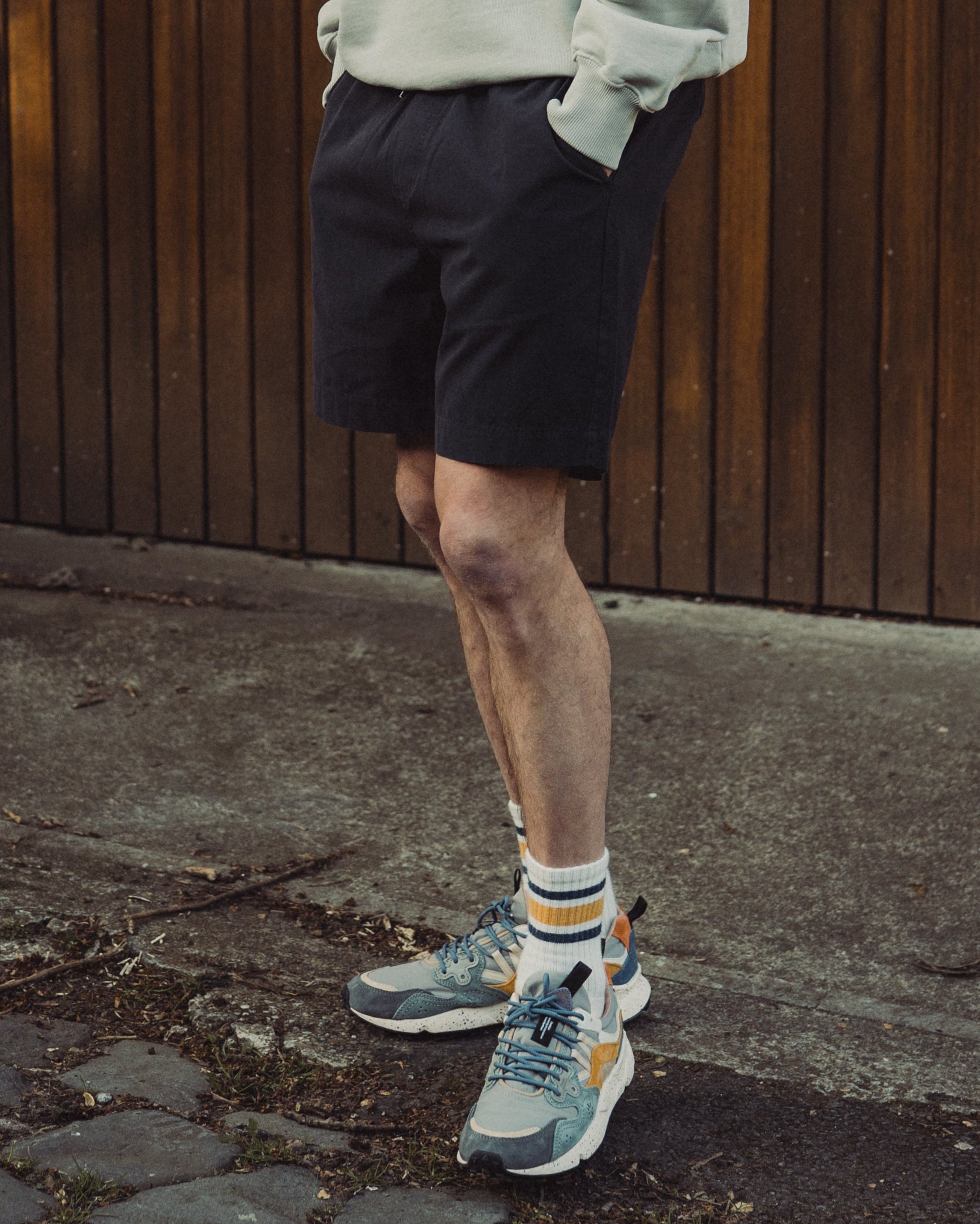 Man wearing a light gray sweatshirt and black shorts standing against a wooden wall.