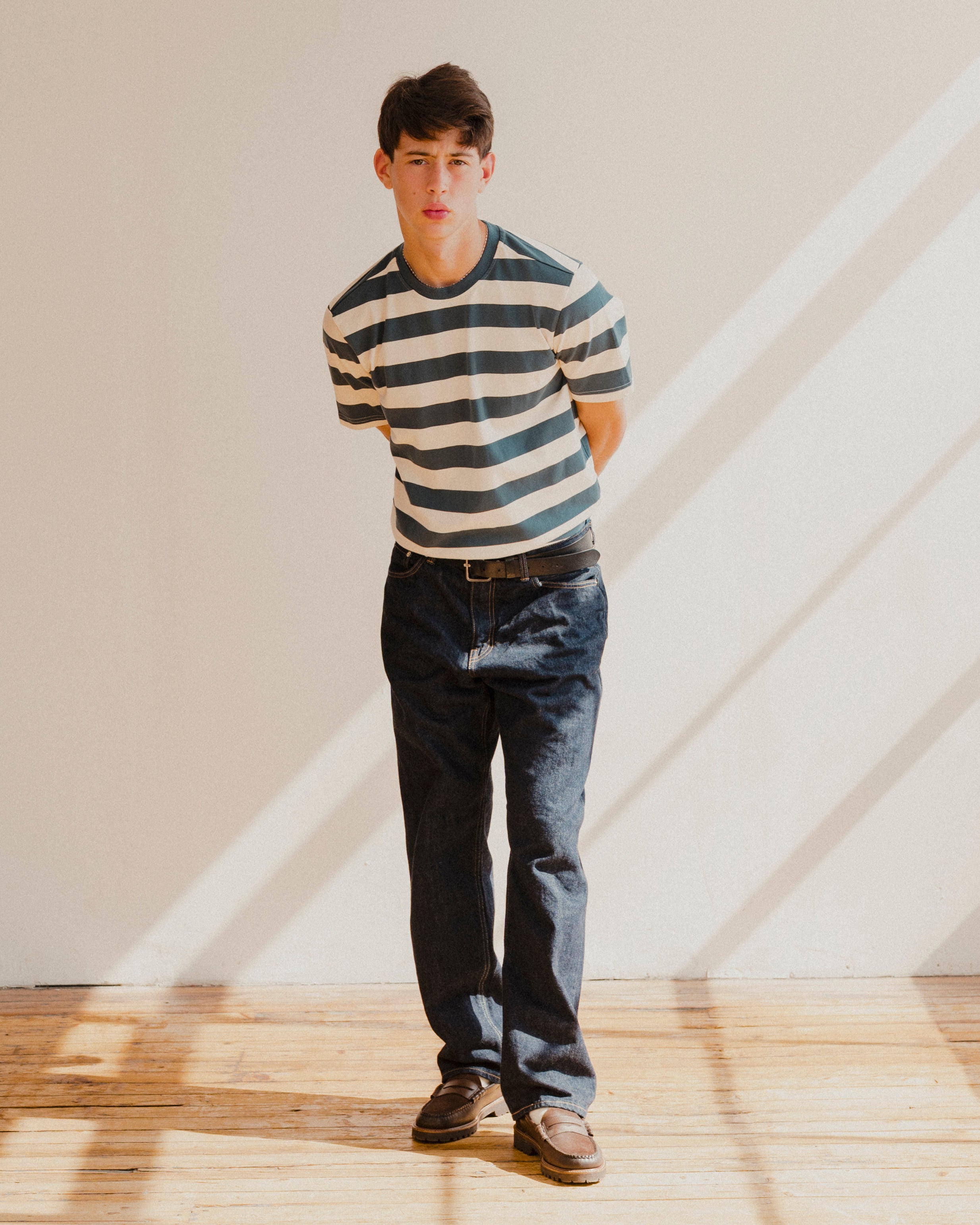 Man wearing a blue and white striped t-shirt and jeans standing in a room with wooden floor and white walls.