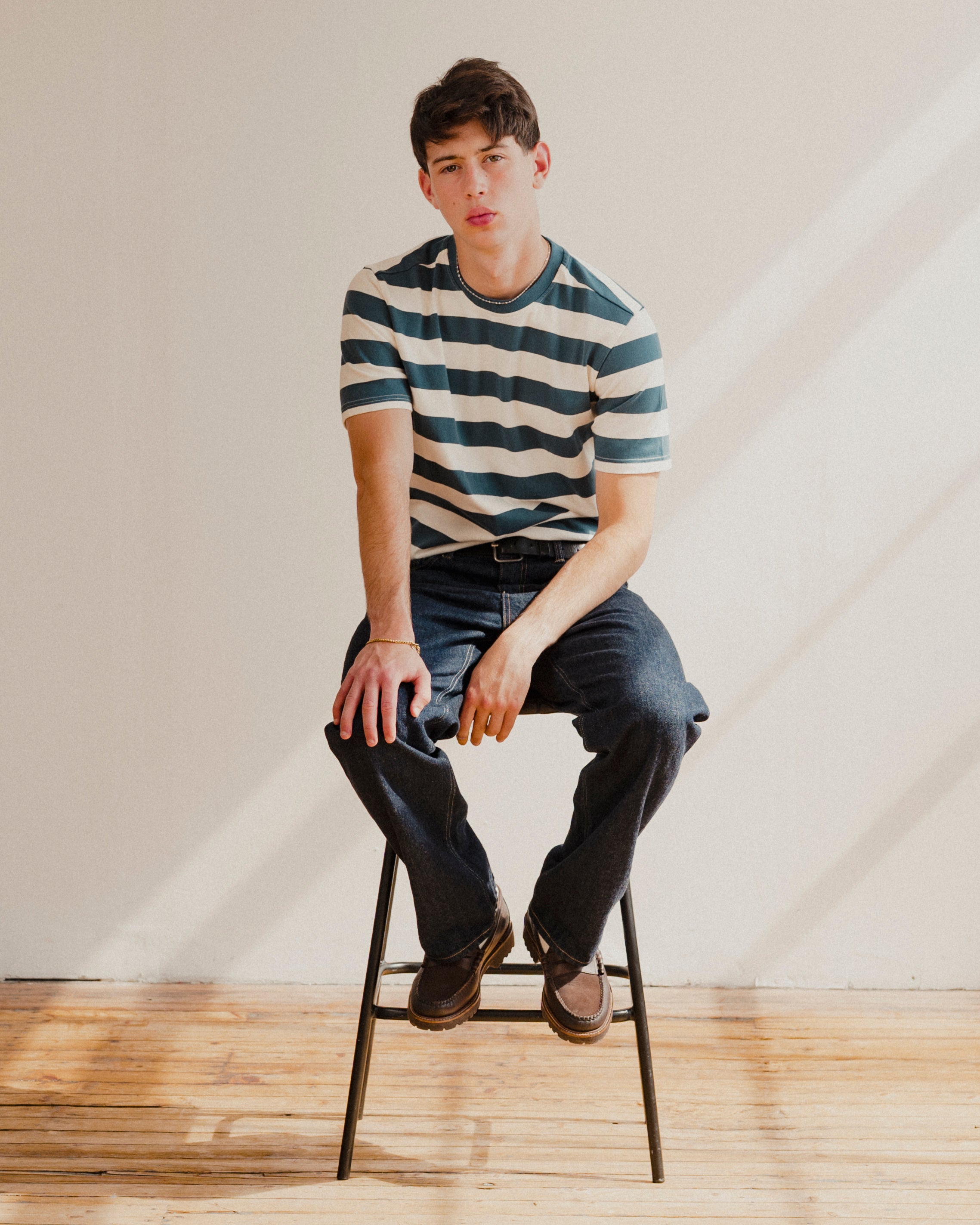 Man sitting on a stool wearing a blue and white striped t-shirt and jeans in a minimalistic setting.