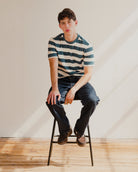 Man sitting on a stool wearing a blue and white striped t-shirt and jeans in a minimalistic setting.