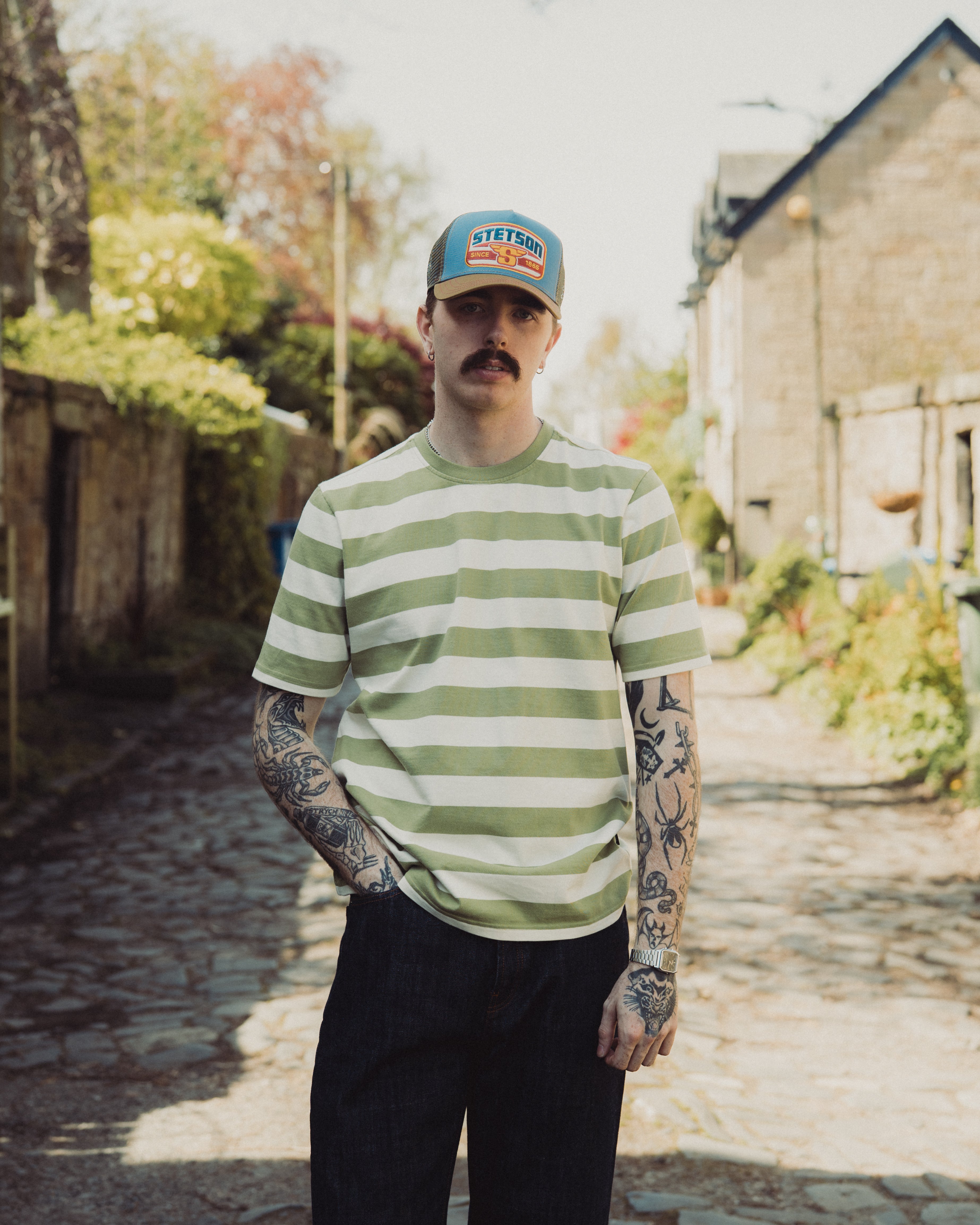 Man wearing a green and white striped shirt and cap standing in a sunlit alleyway.