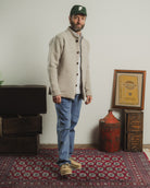 Man wearing a beige cardigan and blue jeans standing in a room with a rug, crates, and a framed document.