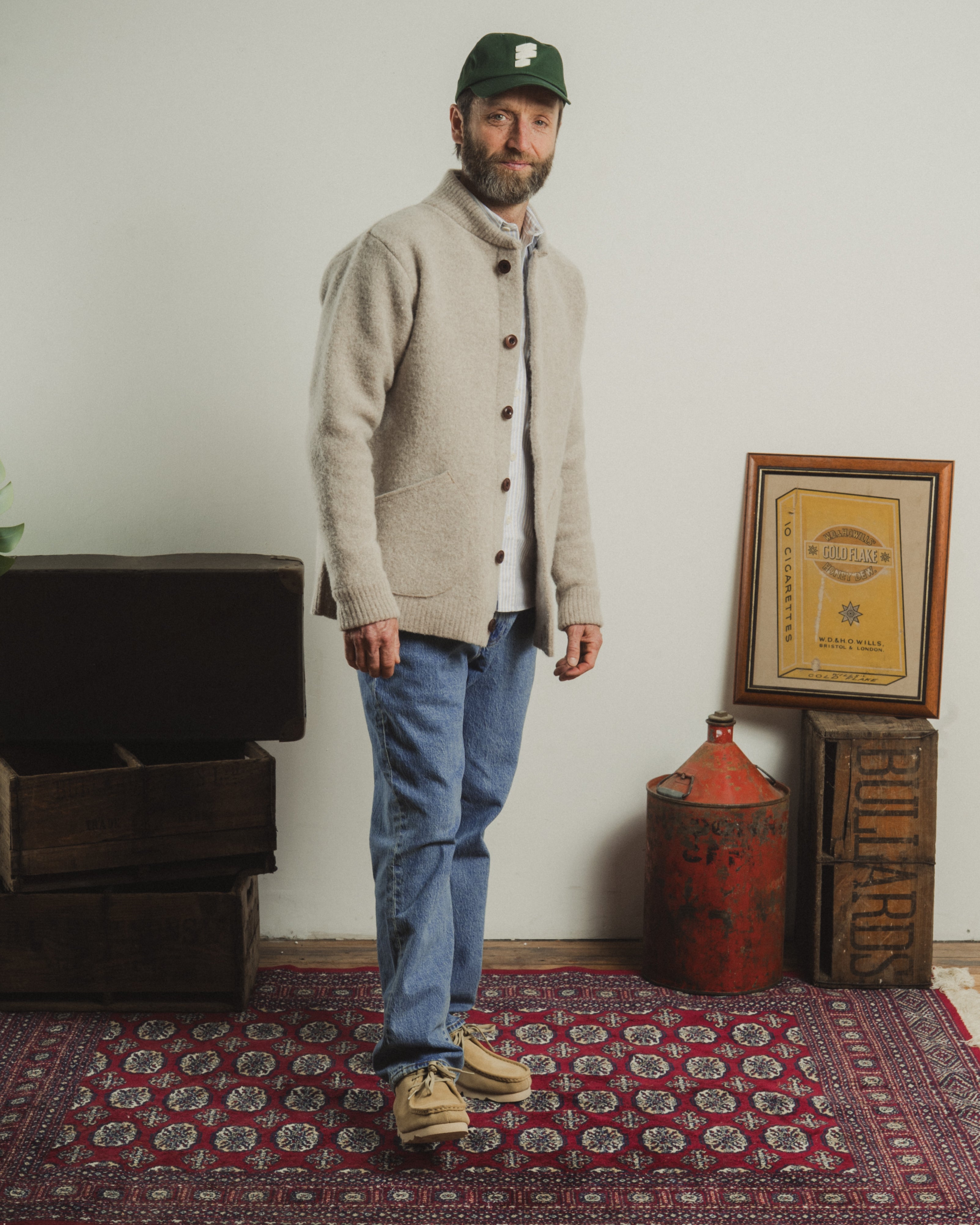 Man wearing a beige cardigan and blue jeans standing in a room with a rug, crates, and a framed document.