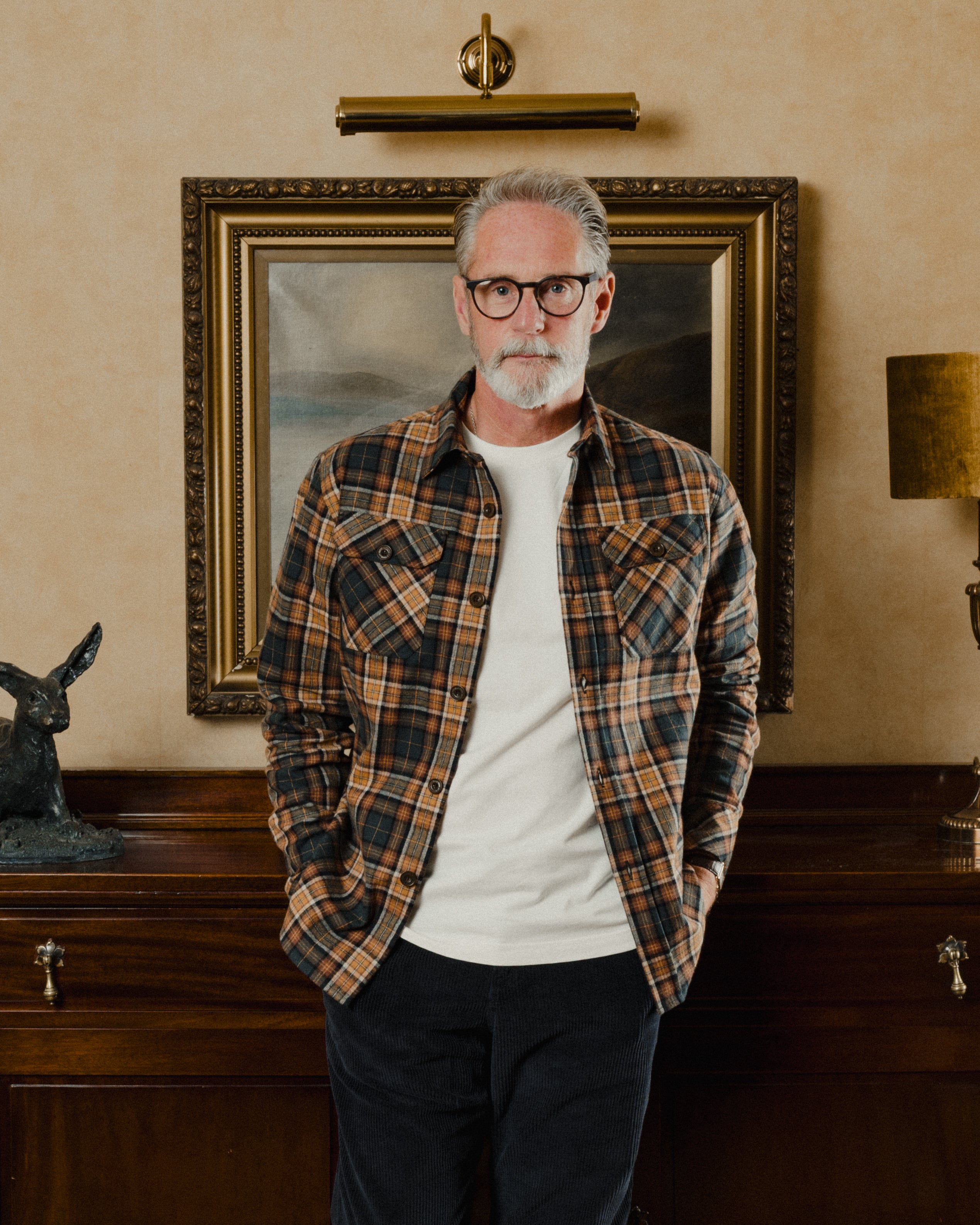 Man wearing a plaid shirt standing in a room with a painting on the wall.