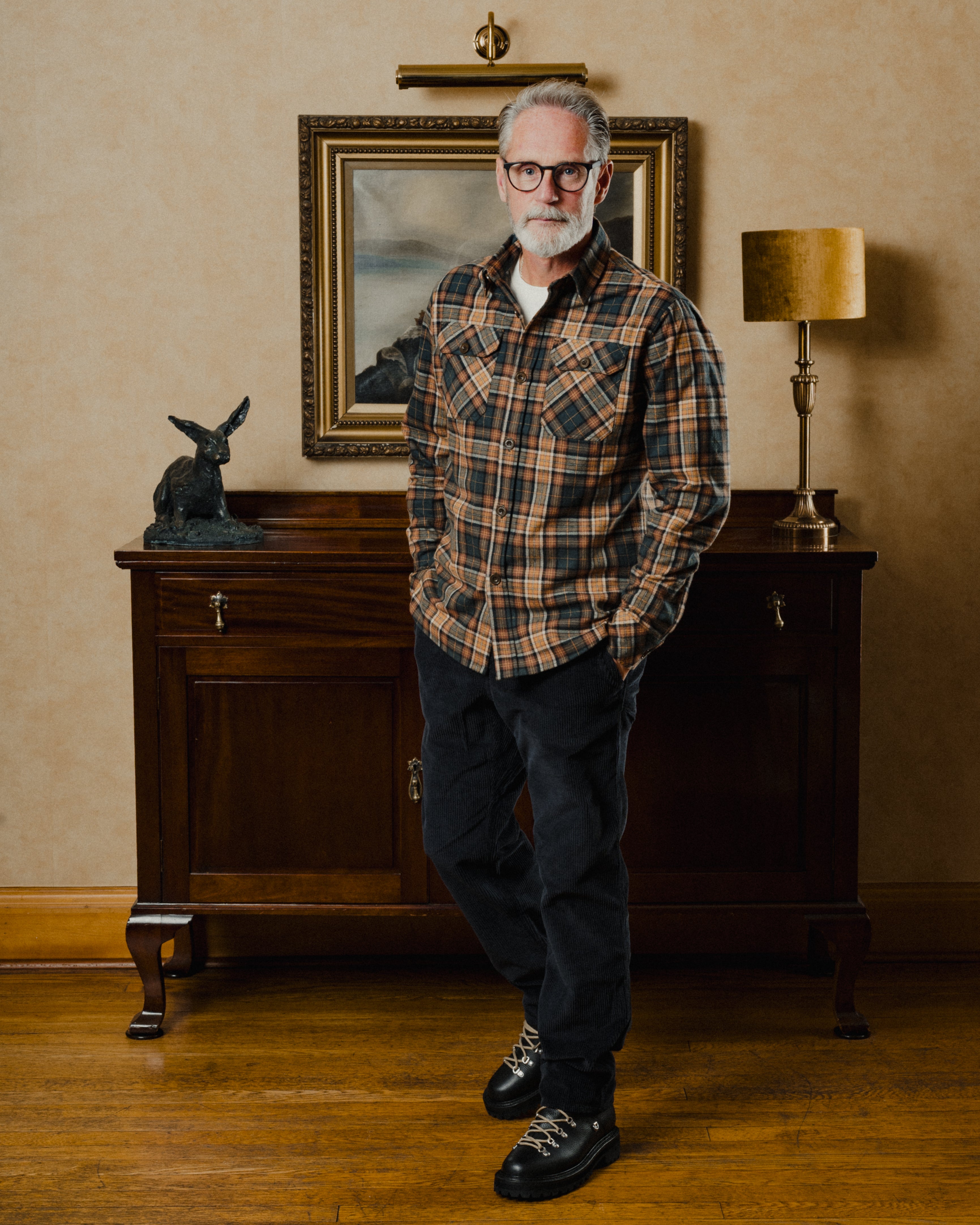 Man wearing a plaid shirt standing in a room with a wooden cabinet and lamp.