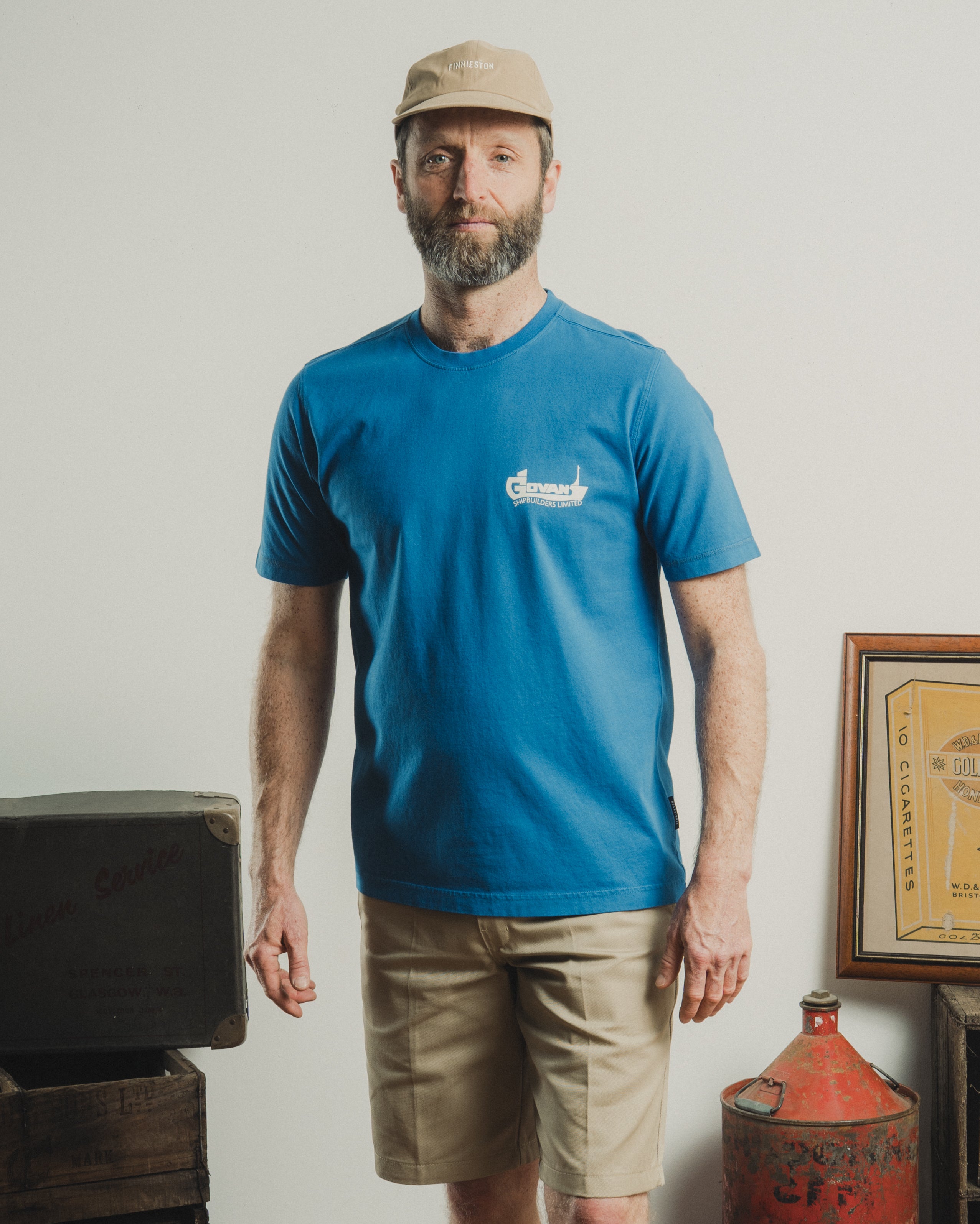 Man wearing a blue t-shirt with a logo, beige cap, and shorts standing in a rustic setting.