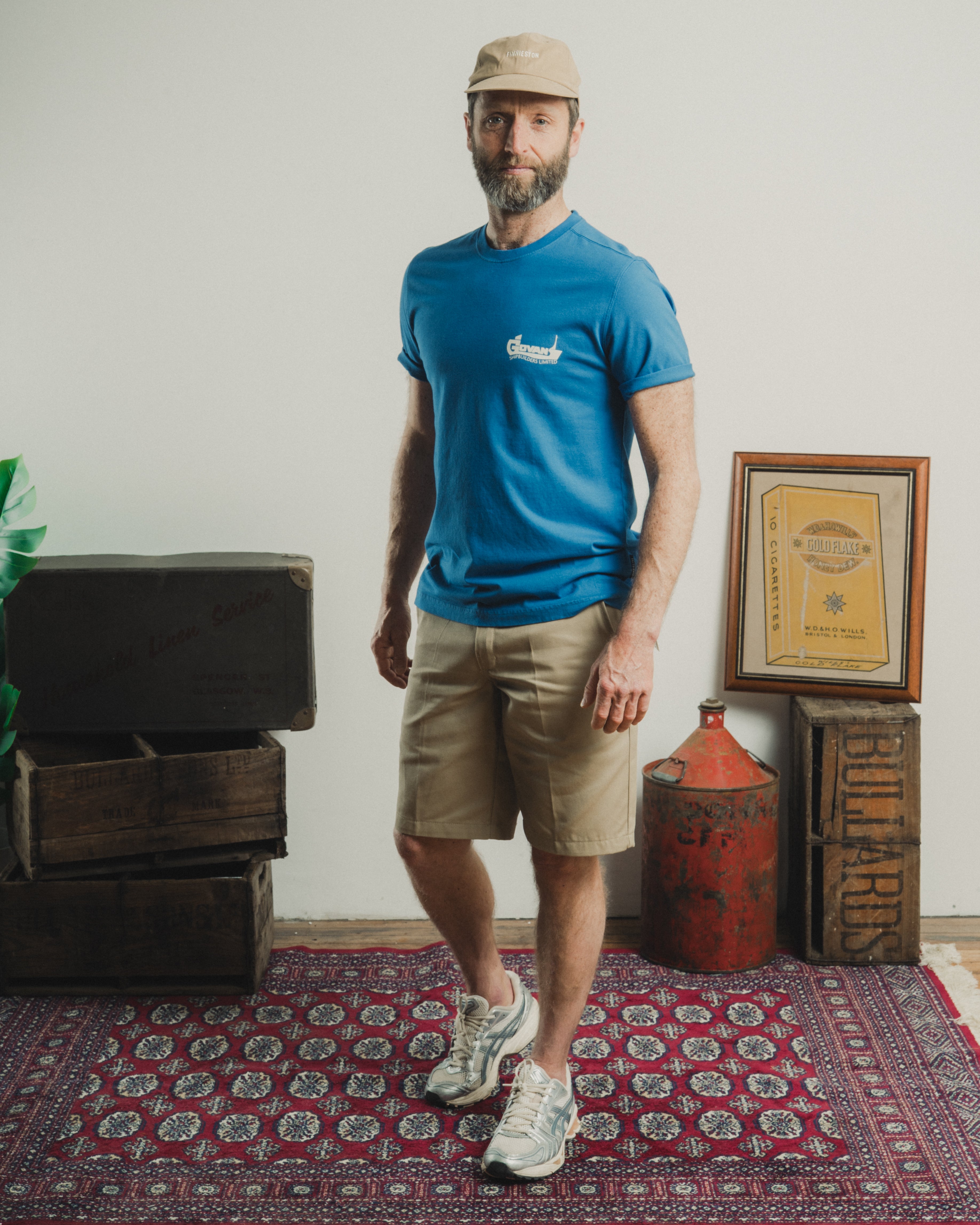 Man wearing a blue t-shirt and beige shorts standing in a room with a rug and wooden crates.