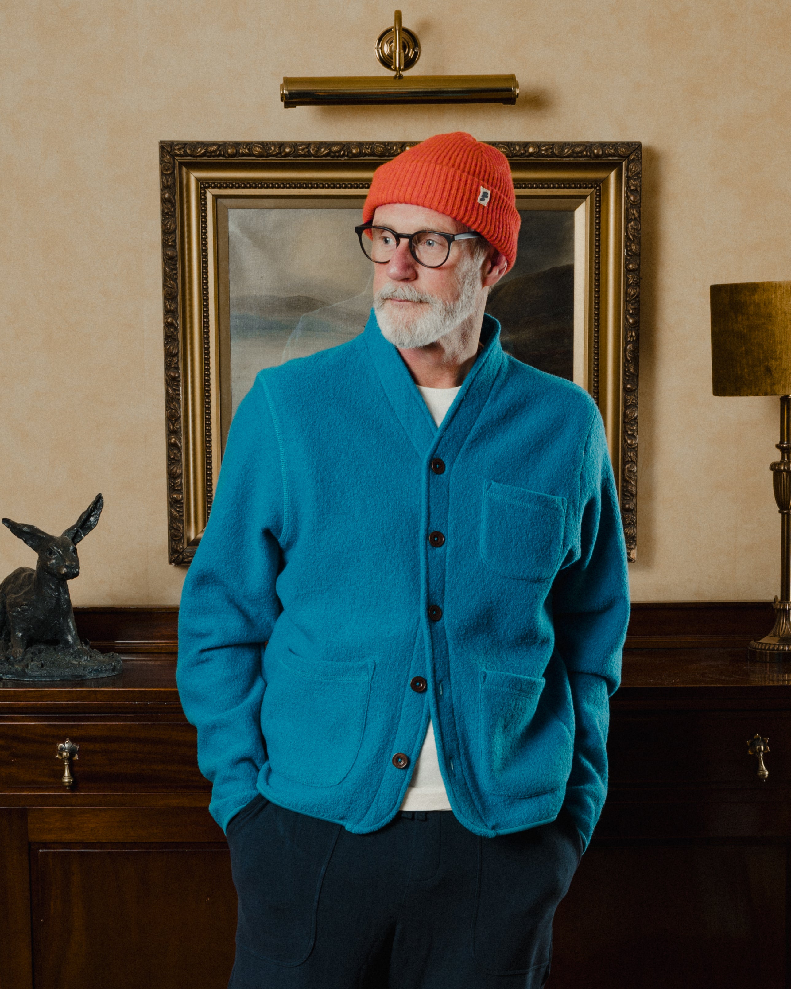 Man wearing a blue cardigan and red beanie standing in a room with a painting and lamp.