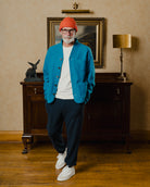 Man wearing a blue jacket, white shirt, navy pants, and orange beanie standing in a room with a lamp and painting.