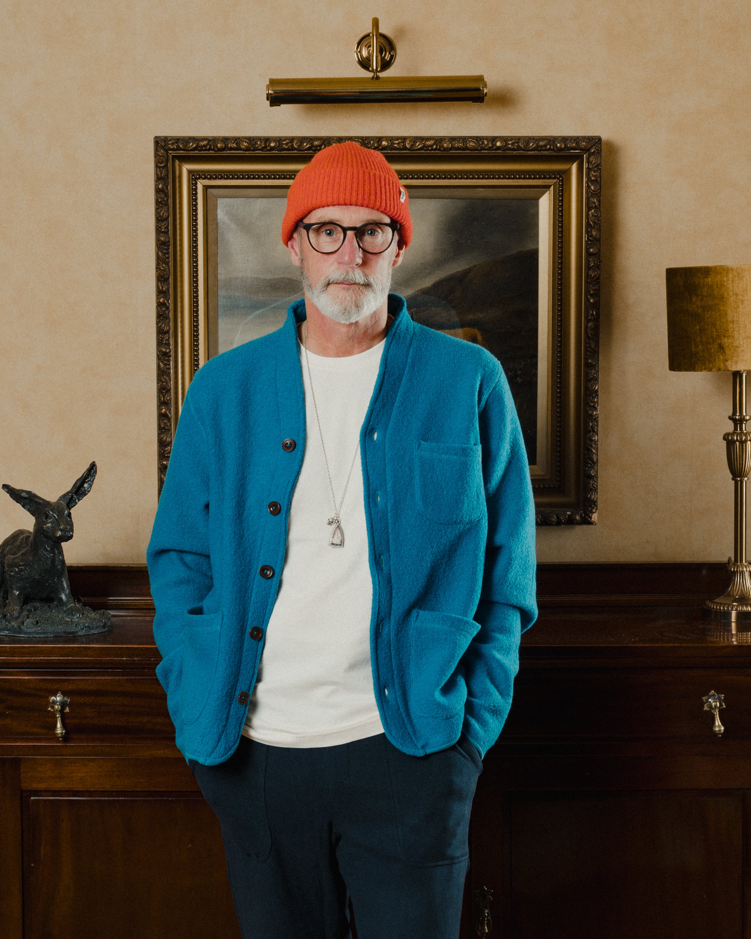 Man wearing a blue jacket and red beanie standing in a room with a painting and lamp.