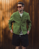 Man wearing a green cardigan, sunglasses, and black shorts against a wooden background