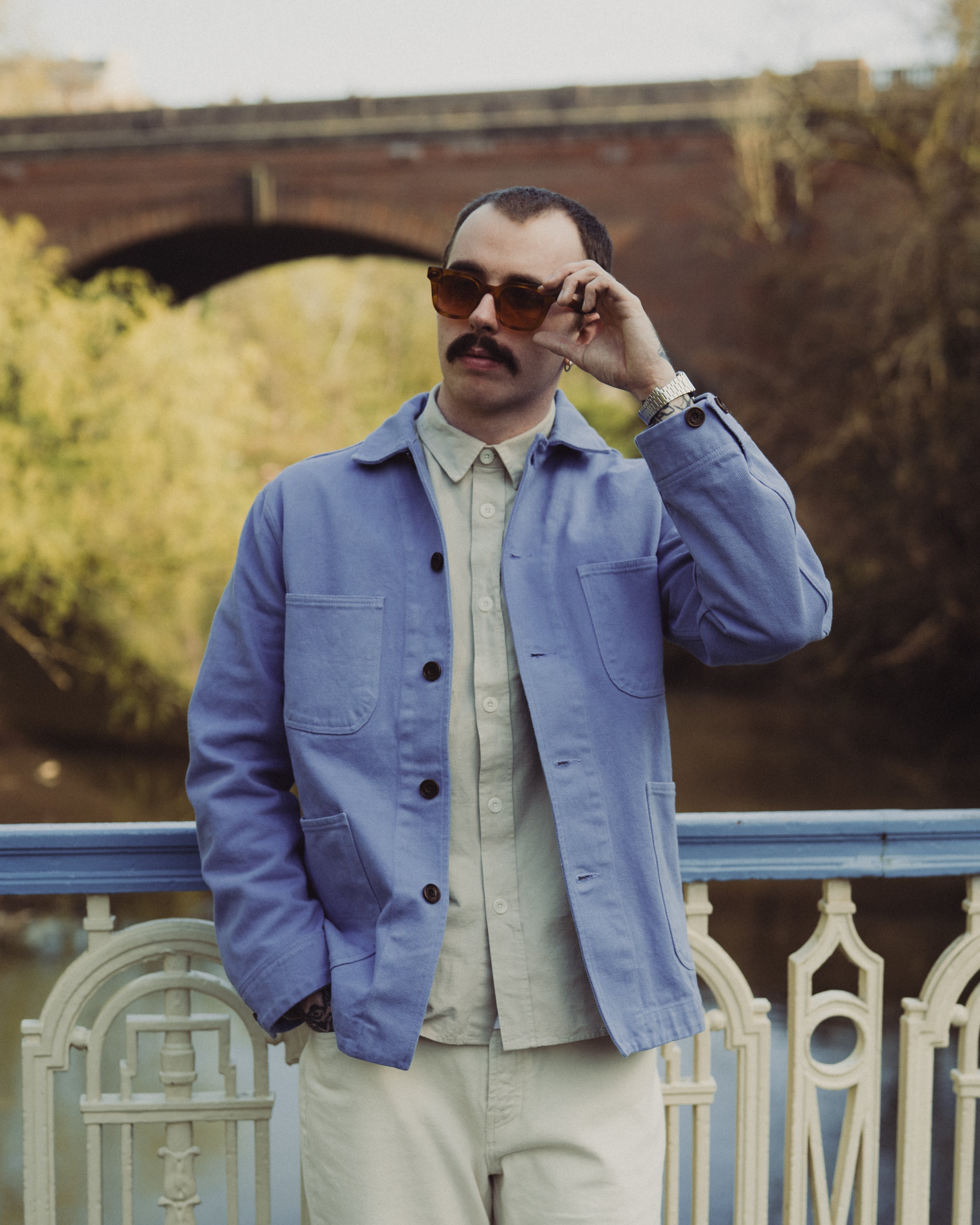 Man wearing a blue jacket and sunglasses, standing by a railing with a bridge in the background.