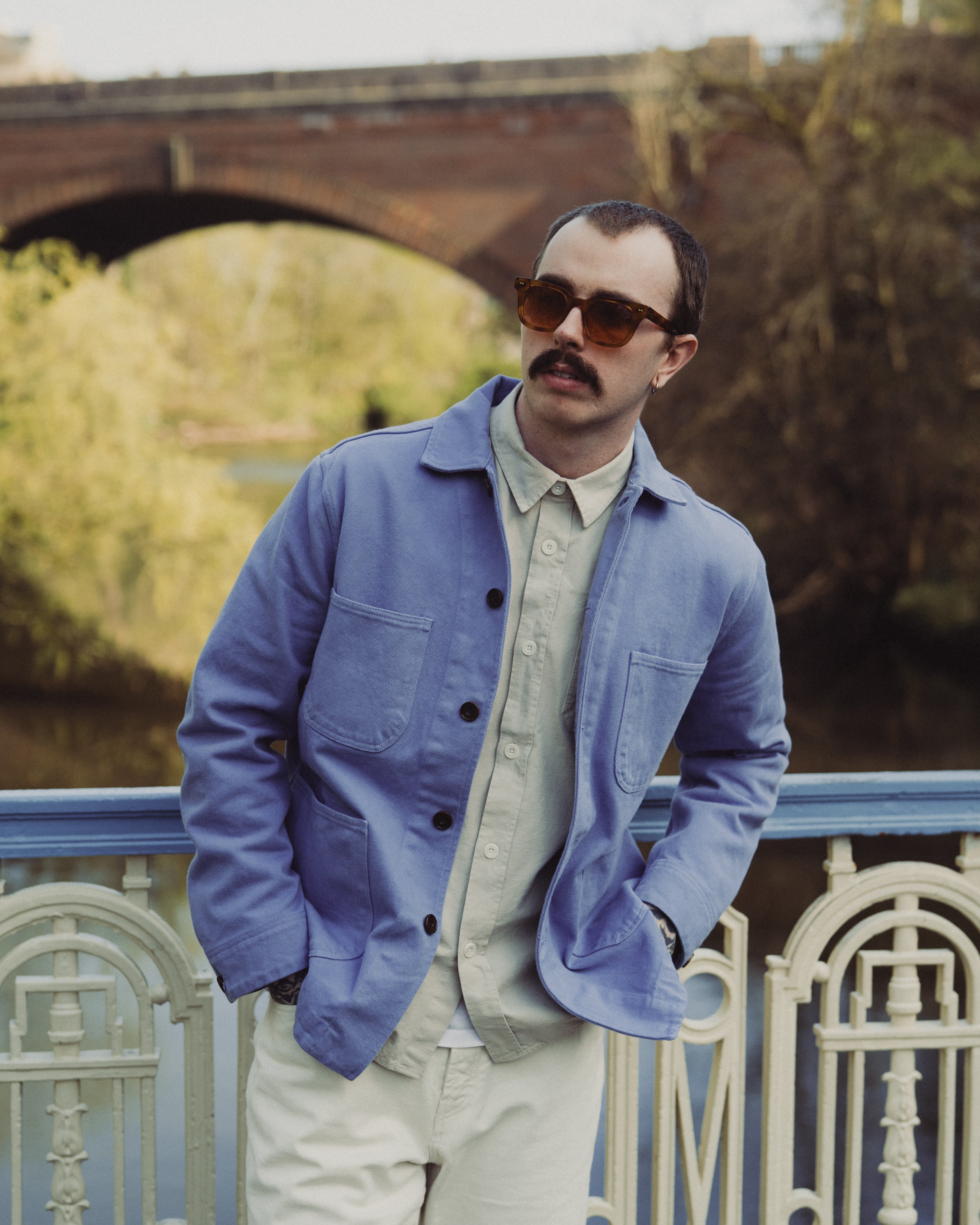 Man wearing a blue jacket and sunglasses standing by a railing with a bridge and trees in the background