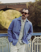 Man wearing a blue jacket and sunglasses standing by a railing with a bridge and trees in the background