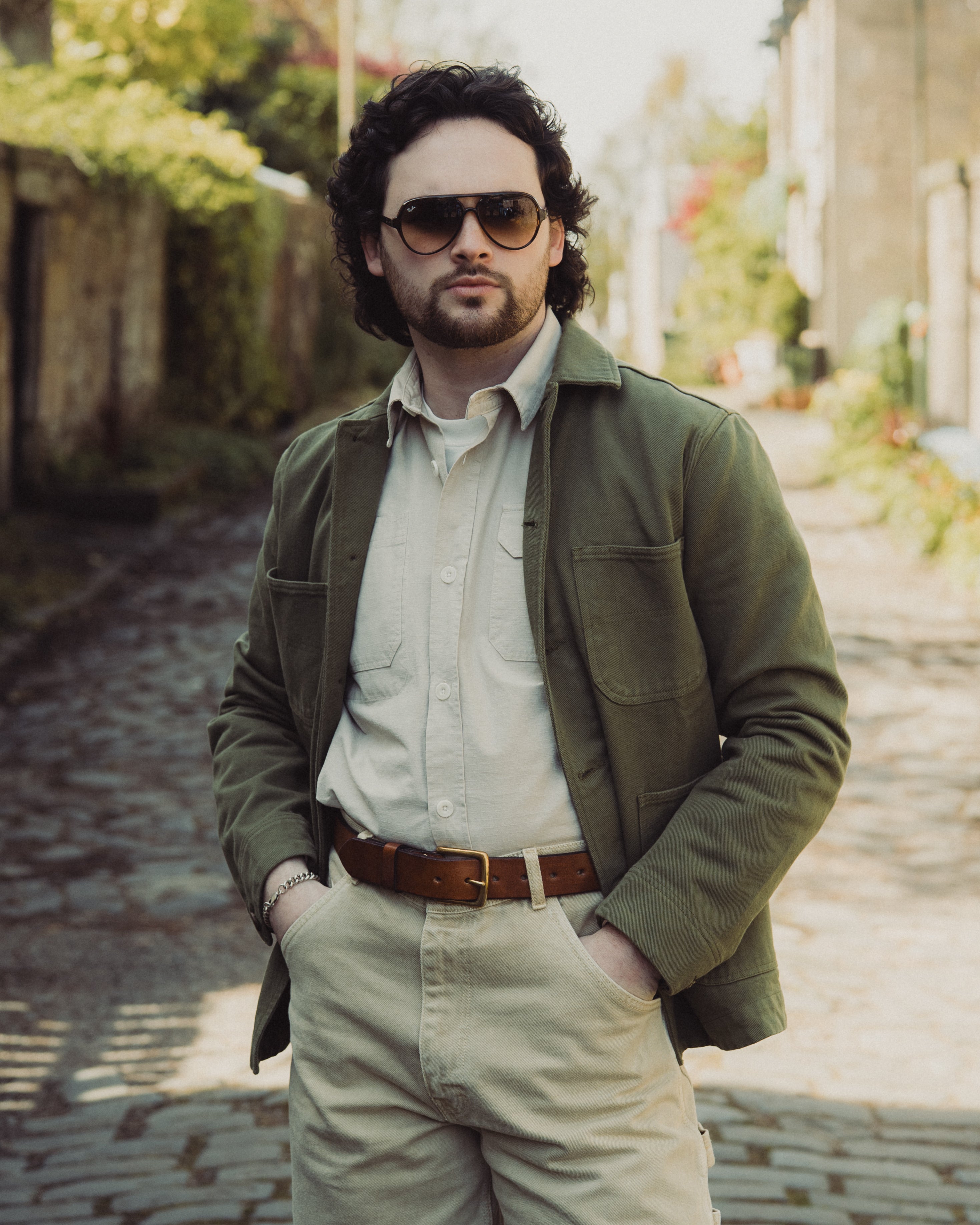Man wearing sunglasses and a green jacket standing on a stone path with blurred background