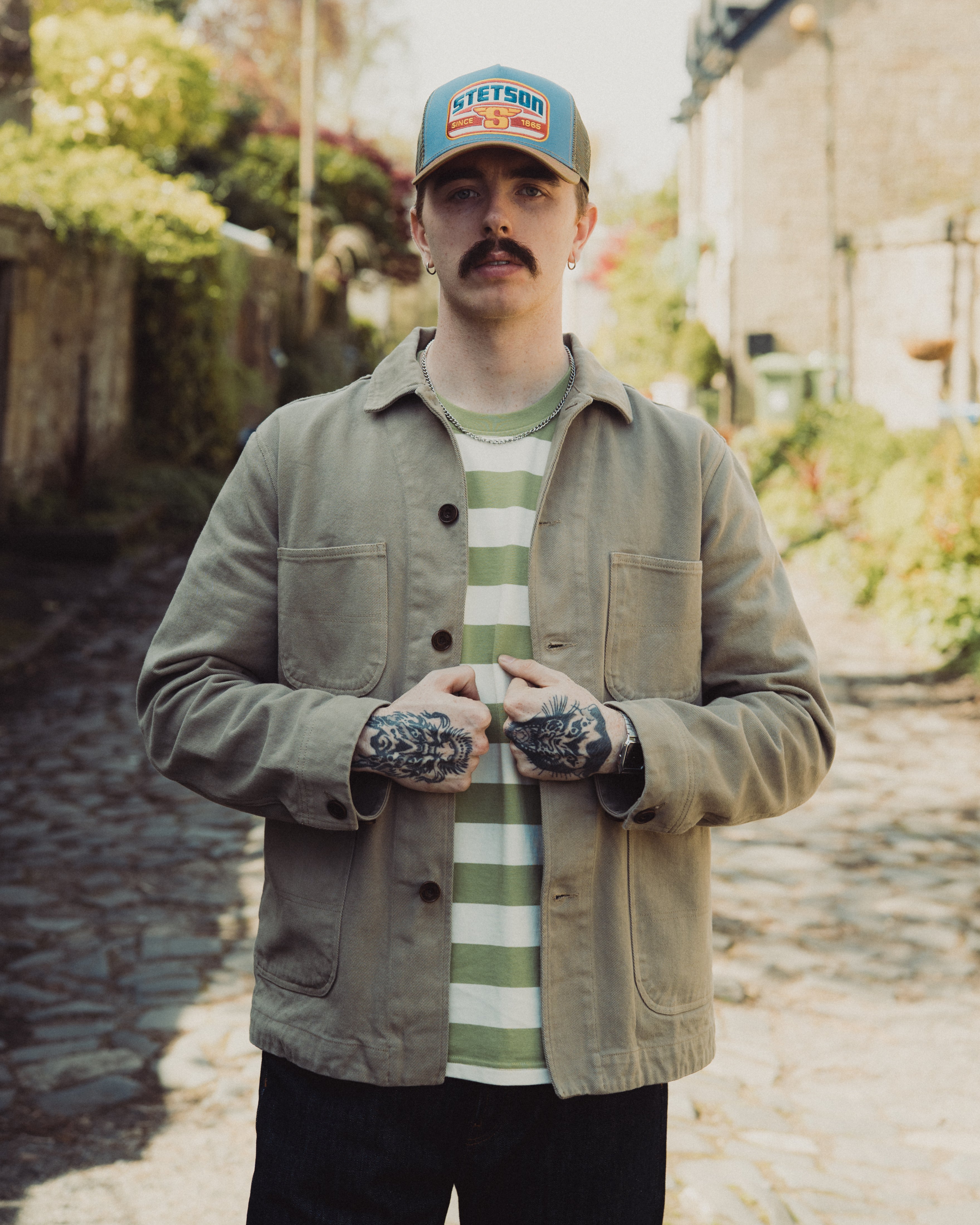 Man wearing a cap and jacket standing on a stone path with greenery