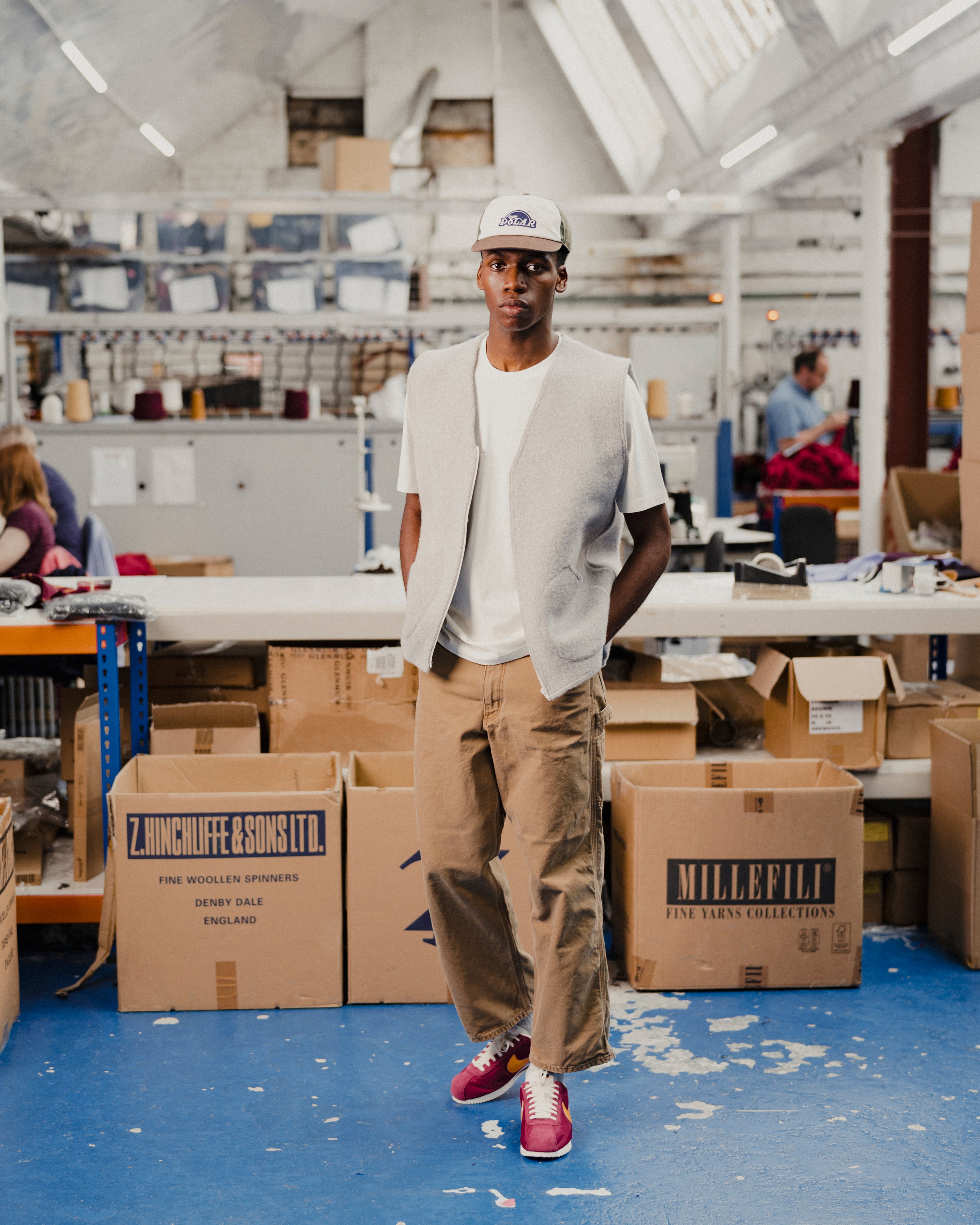 Person wearing a grey wool vest and white t-shirt standing in a warehouse with boxes and workstations in the background