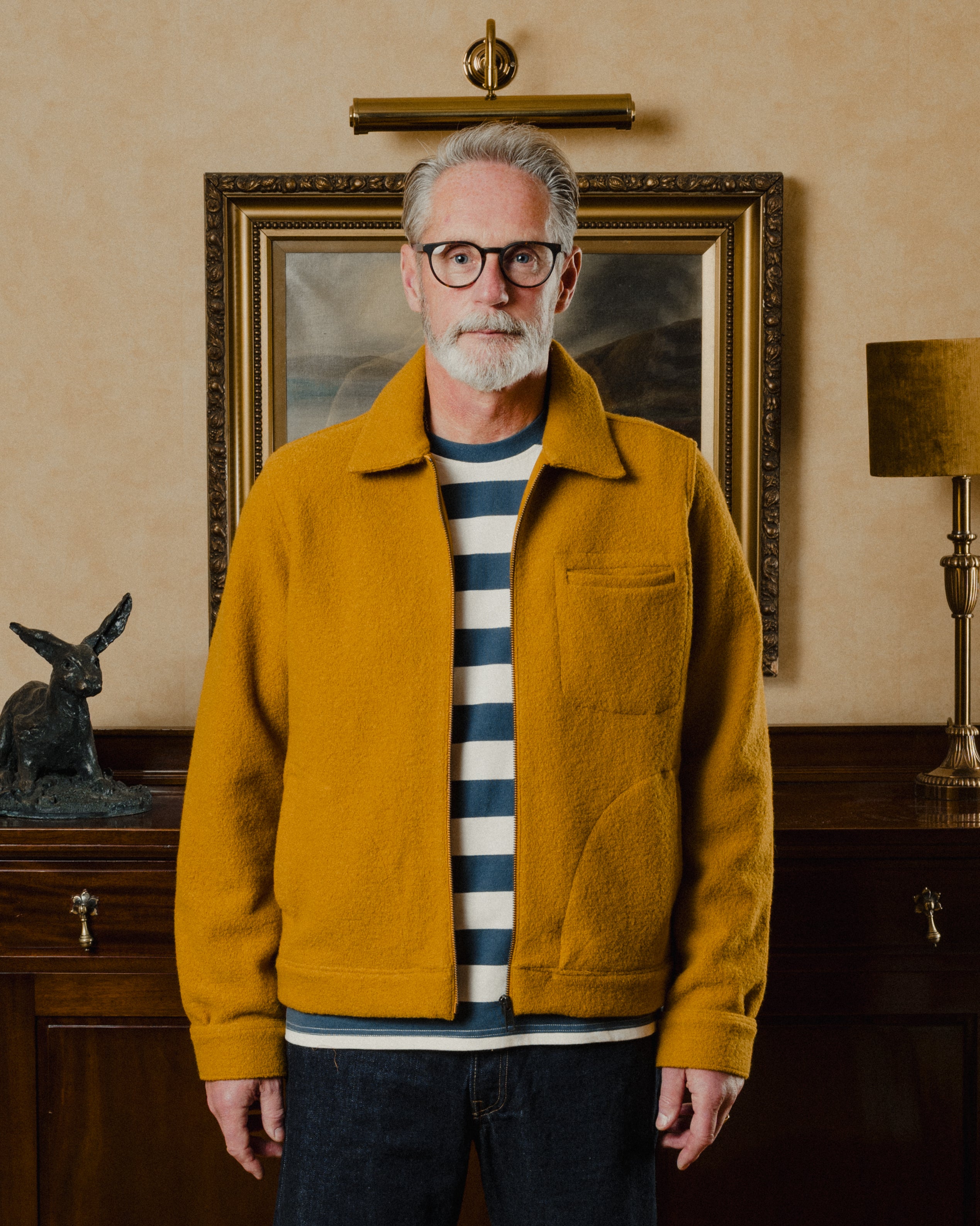 Man wearing a mustard yellow jacket in a room with a painting and lamp in the background