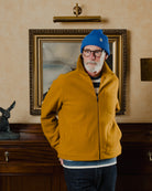 Man wearing a yellow jacket and blue beanie in a room with a painting on the wall.
