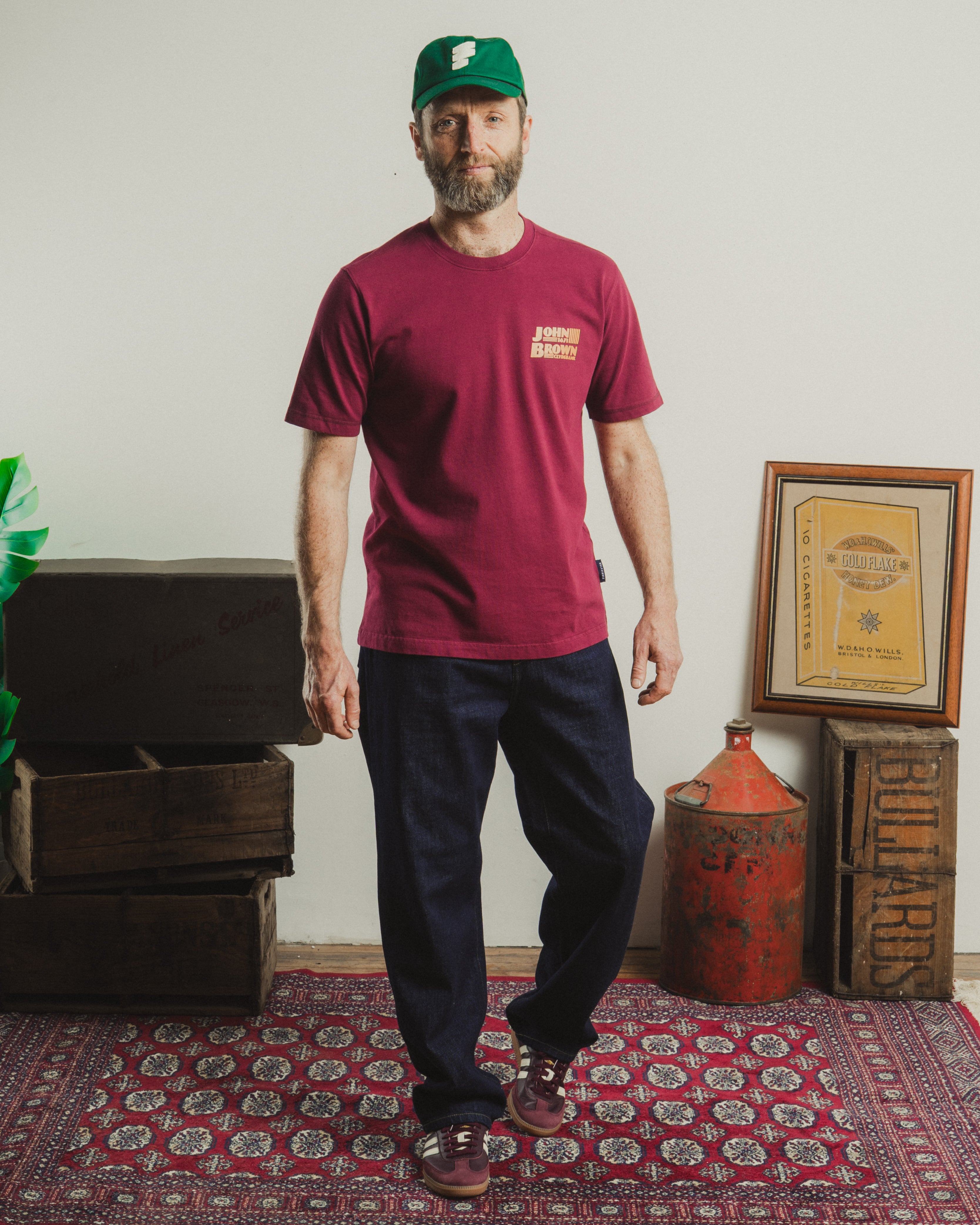 Man wearing a red t-shirt and green cap standing in a room with a rug, boxes, and a framed document.