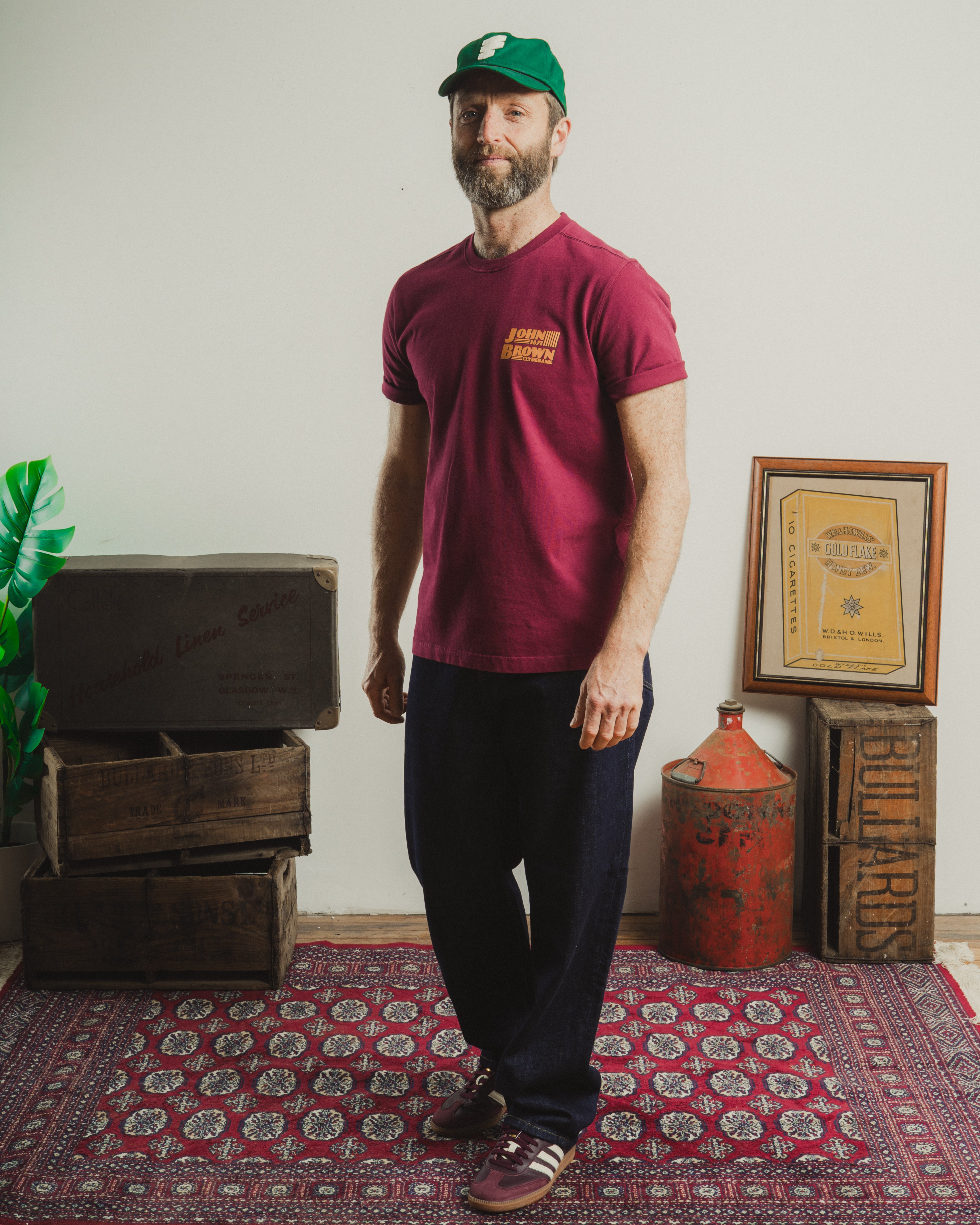 Man wearing a maroon t-shirt with a logo, standing in a room with wooden crates and a rug.