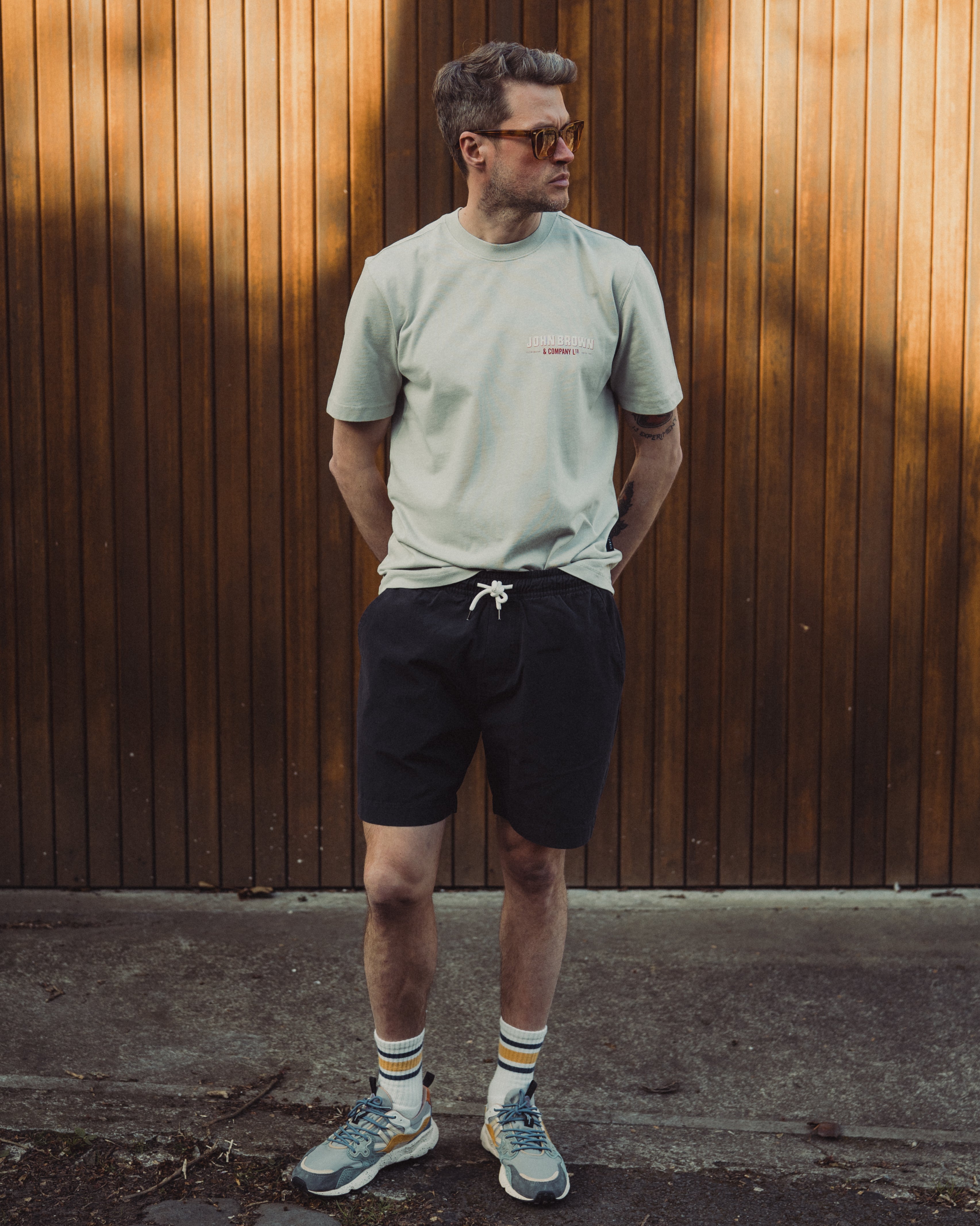 Man standing against a wooden wall wearing a light green t-shirt, black shorts, and colorful sneakers.