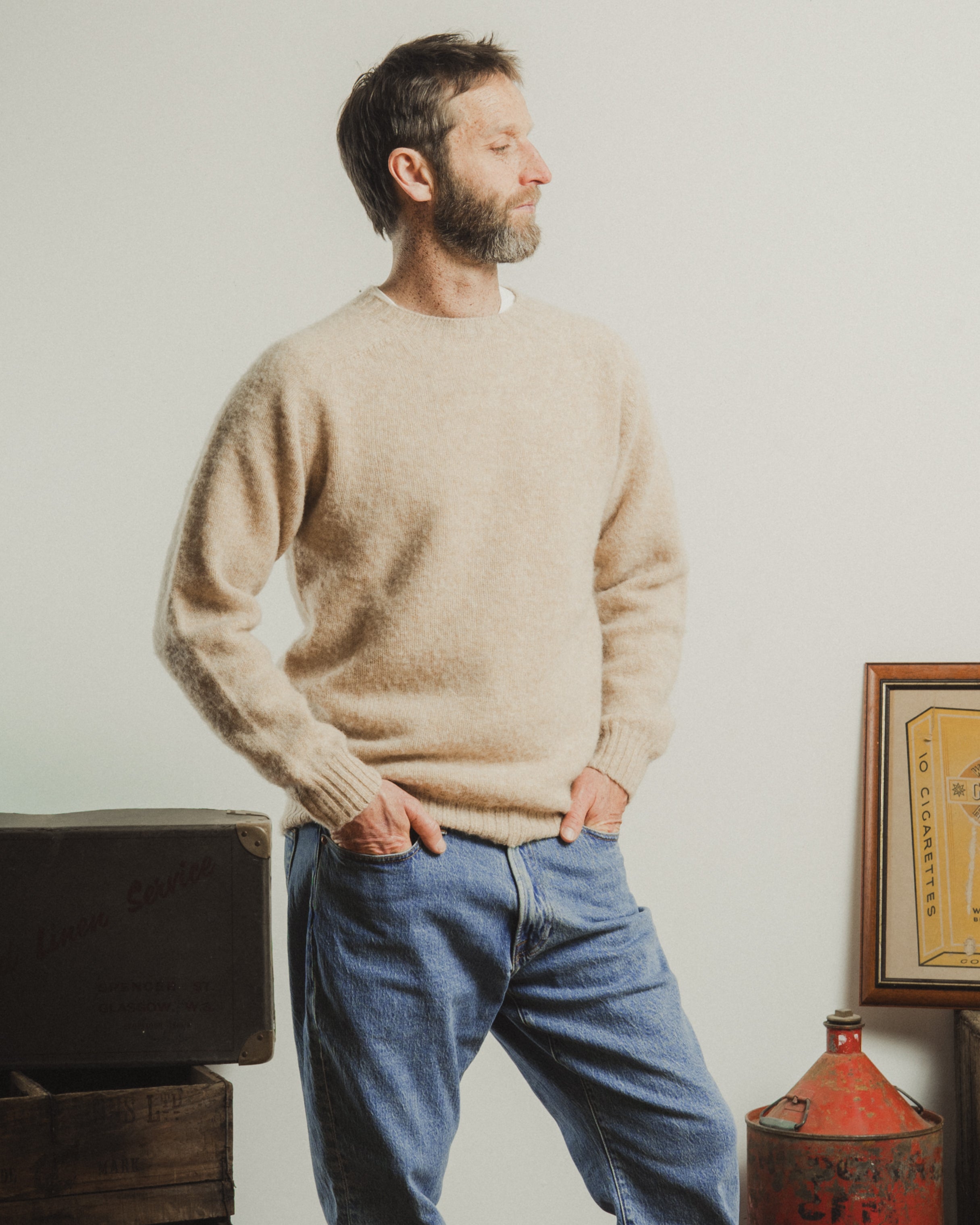 Man wearing a beige sweater and blue jeans standing in a room with a white wall.