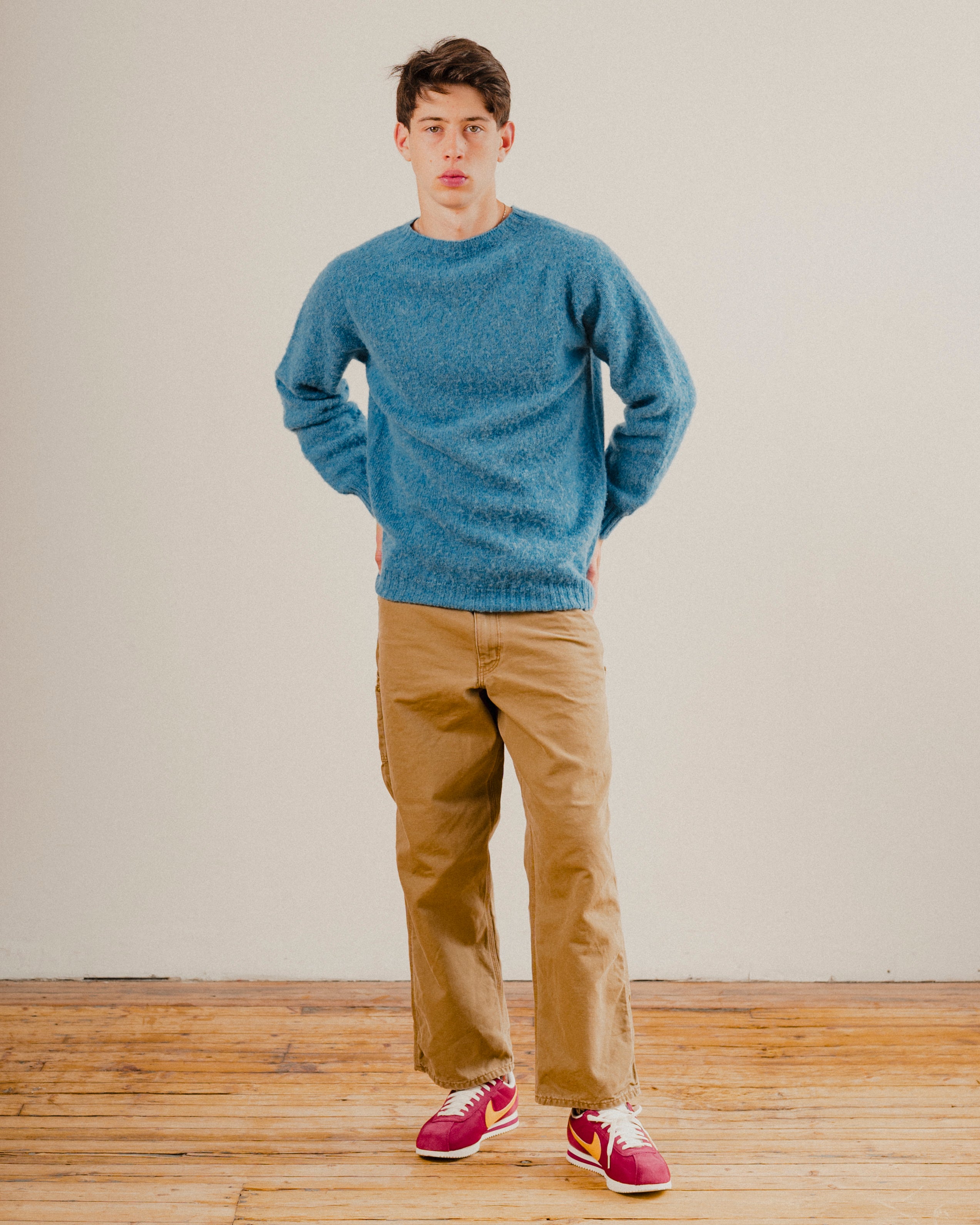 Man in a blue wool knit crewneck jumper sweatshirt on a plain background with a wooden floor
