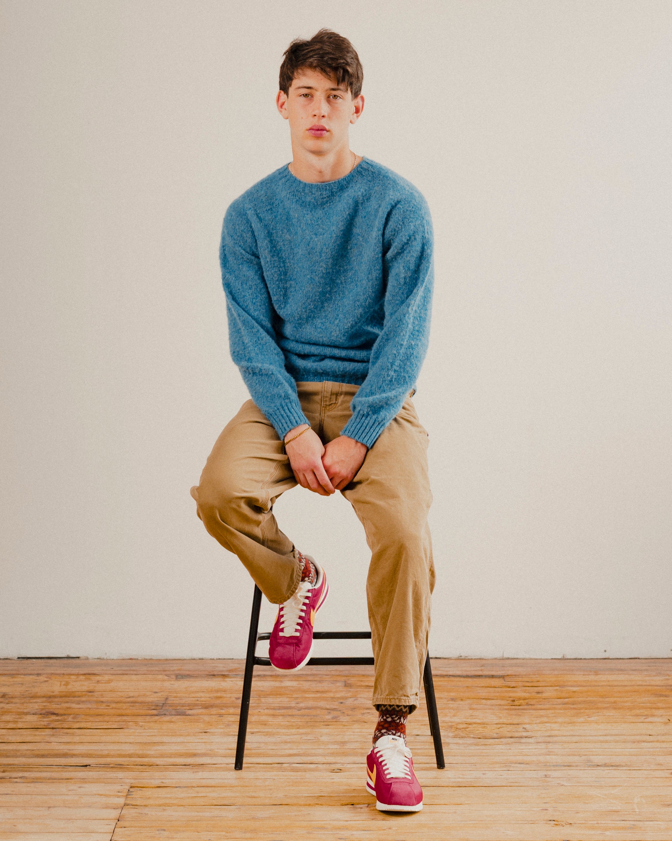 Man sat on stool in a blue wool knit crewneck jumper sweatshirt with a wooden floor and a plain white background