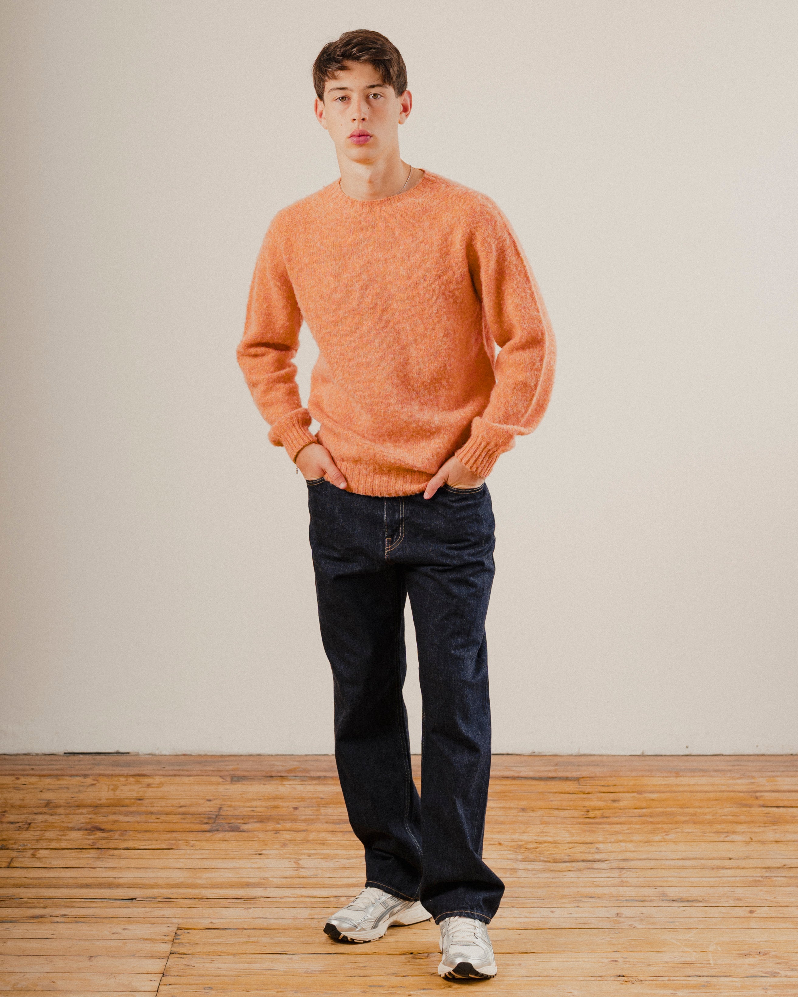 Man wearing orange wool knit sweater with dark blue denim jeans on a wooden floor with a white background