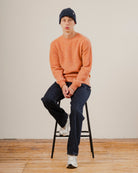 man sat on a stool wearing an orange wool knit crewneck with dark blue denim jeans and a navy blue wool beanie