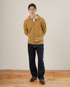 Man wearing a mustard yellow wool cardigan and dark jeans standing against a plain white wall.