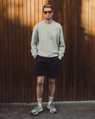 Man wearing a light gray sweatshirt, black shorts, and sunglasses standing against a wooden wall.