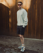 Man wearing a light gray sweatshirt and black shorts standing against a wooden wall.