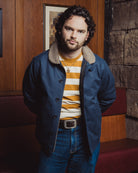 Man wearing a blue coat with a fur-lined hood, striped sweater, and jeans in an indoor setting.