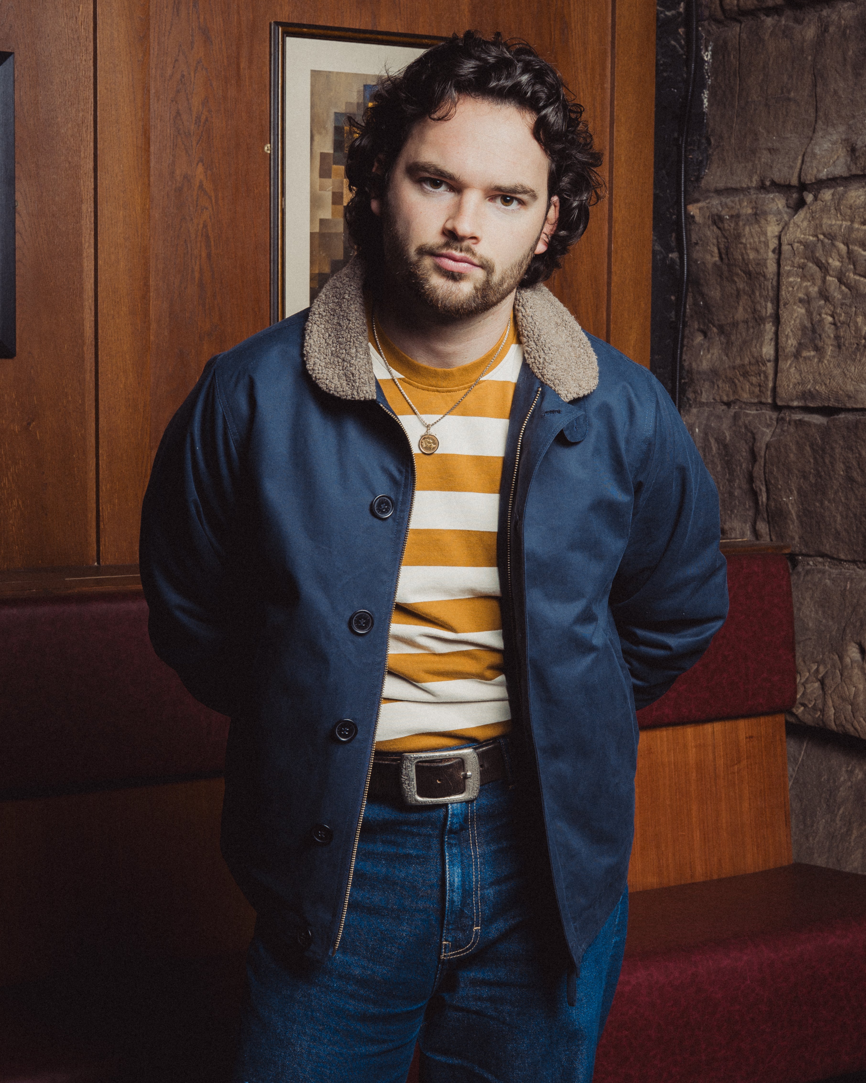 Man wearing a blue coat with a fur-lined hood, striped sweater, and jeans in an indoor setting.