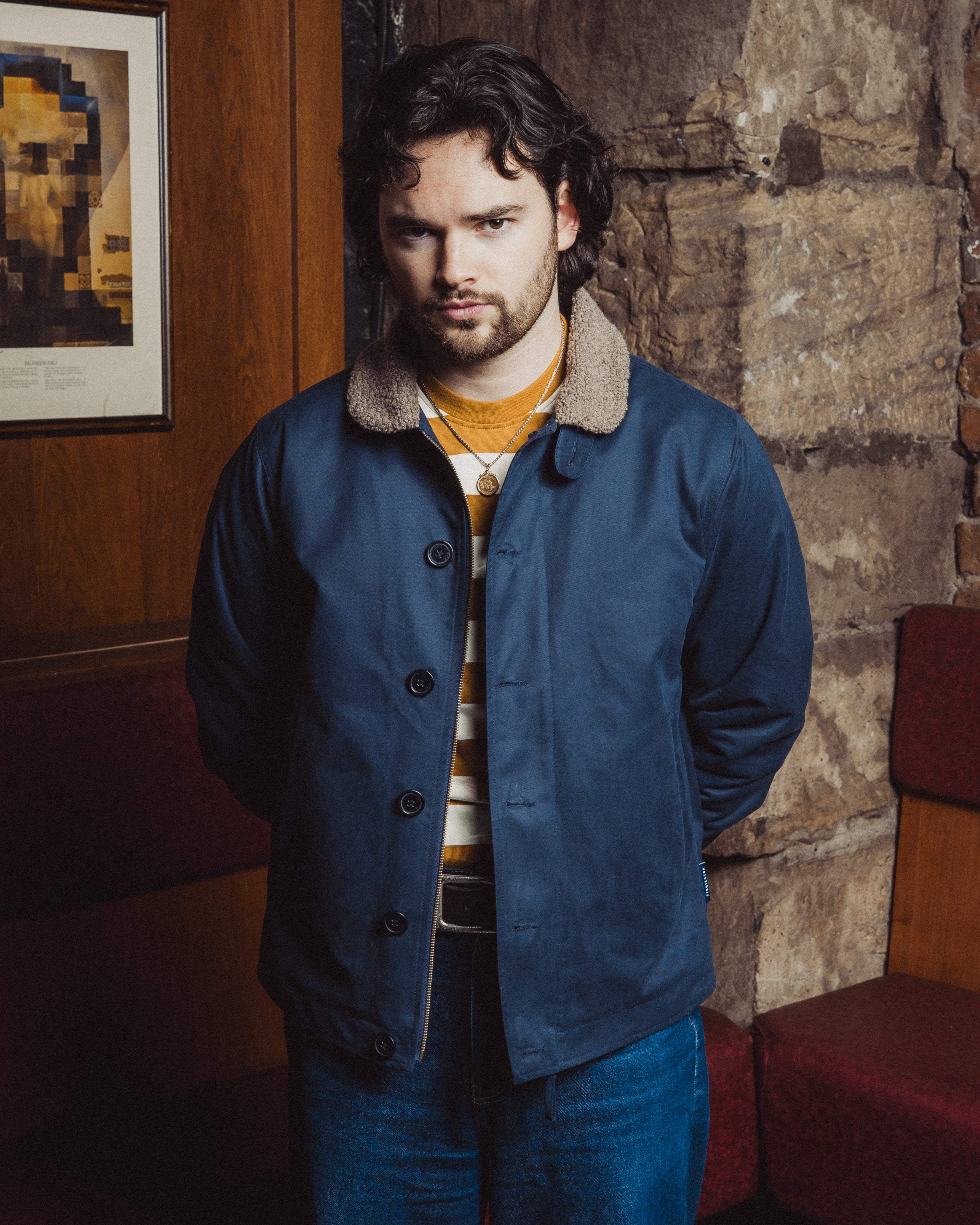 Man wearing a blue coat with a fur-lined hood in a room with stone walls and a framed picture.