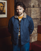 Man wearing a blue coat with a fur-lined hood in a room with stone walls and a framed picture.