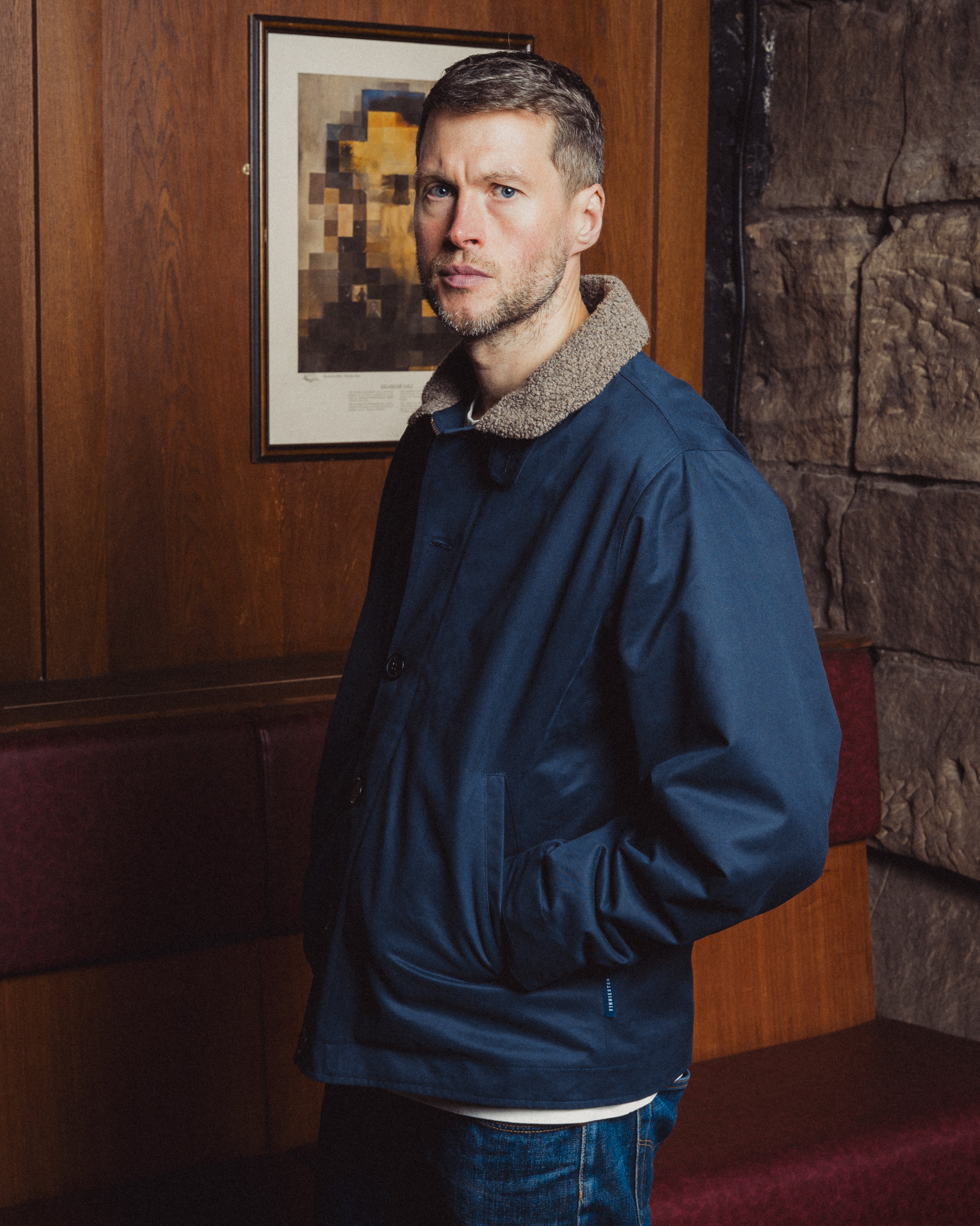 Man wearing a blue jacket in a room with wooden paneling and a painting on the wall.