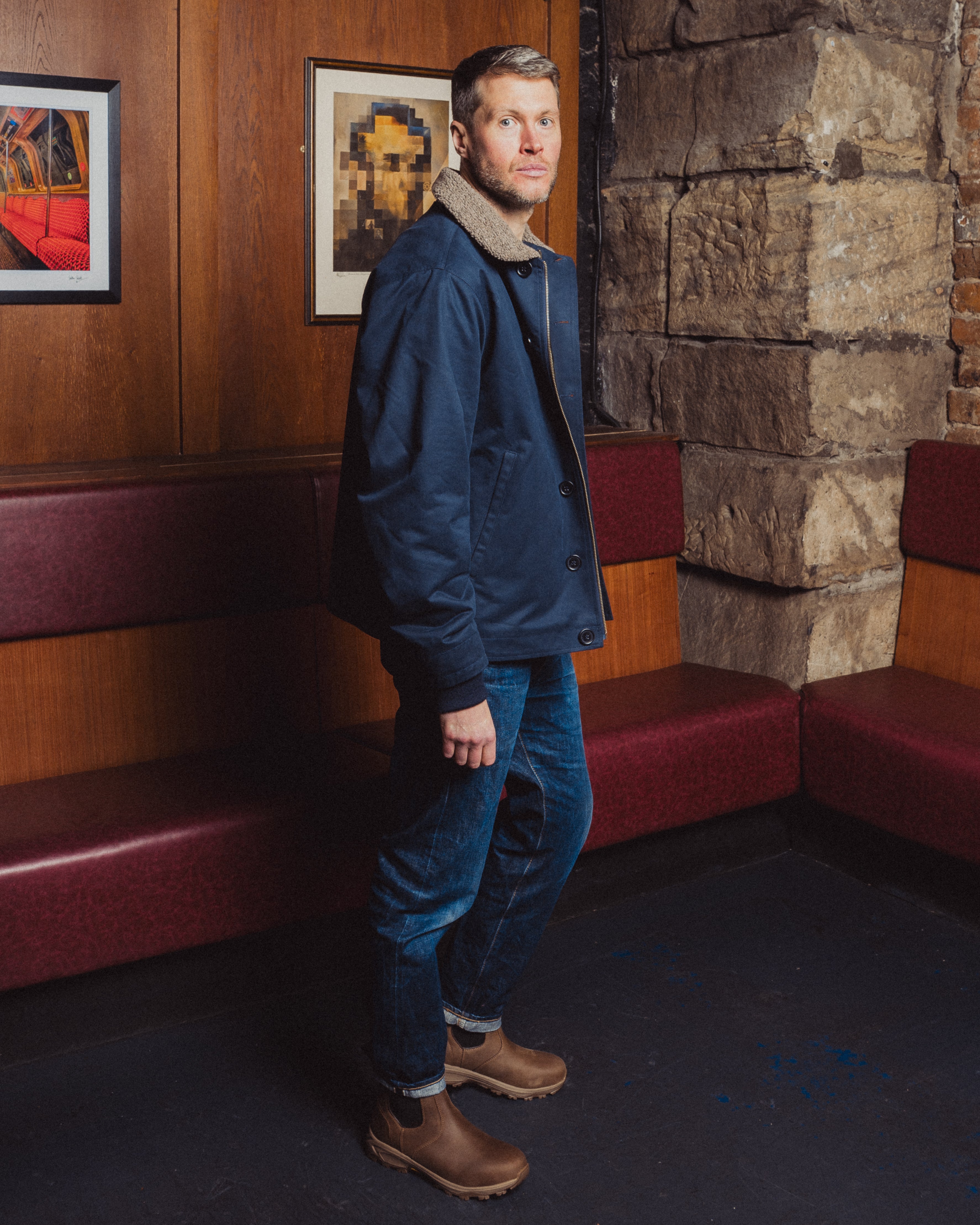 Man in a blue jacket standing in a room with wooden paneling and framed pictures.