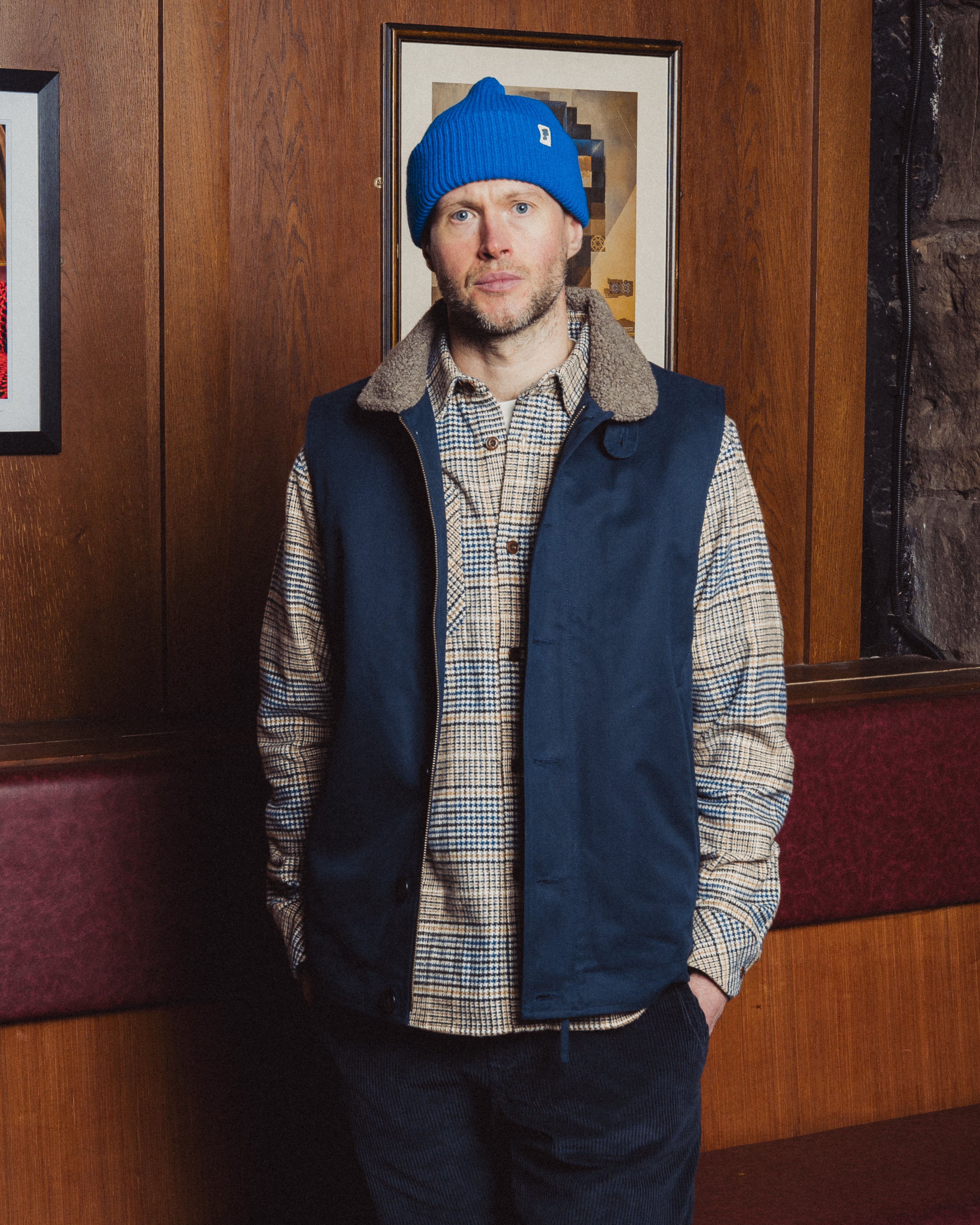 Man wearing a blue beanie and navy vest over a plaid shirt in an indoor setting.