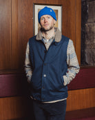 Man wearing a blue vest and blue beanie standing against a wooden wall.