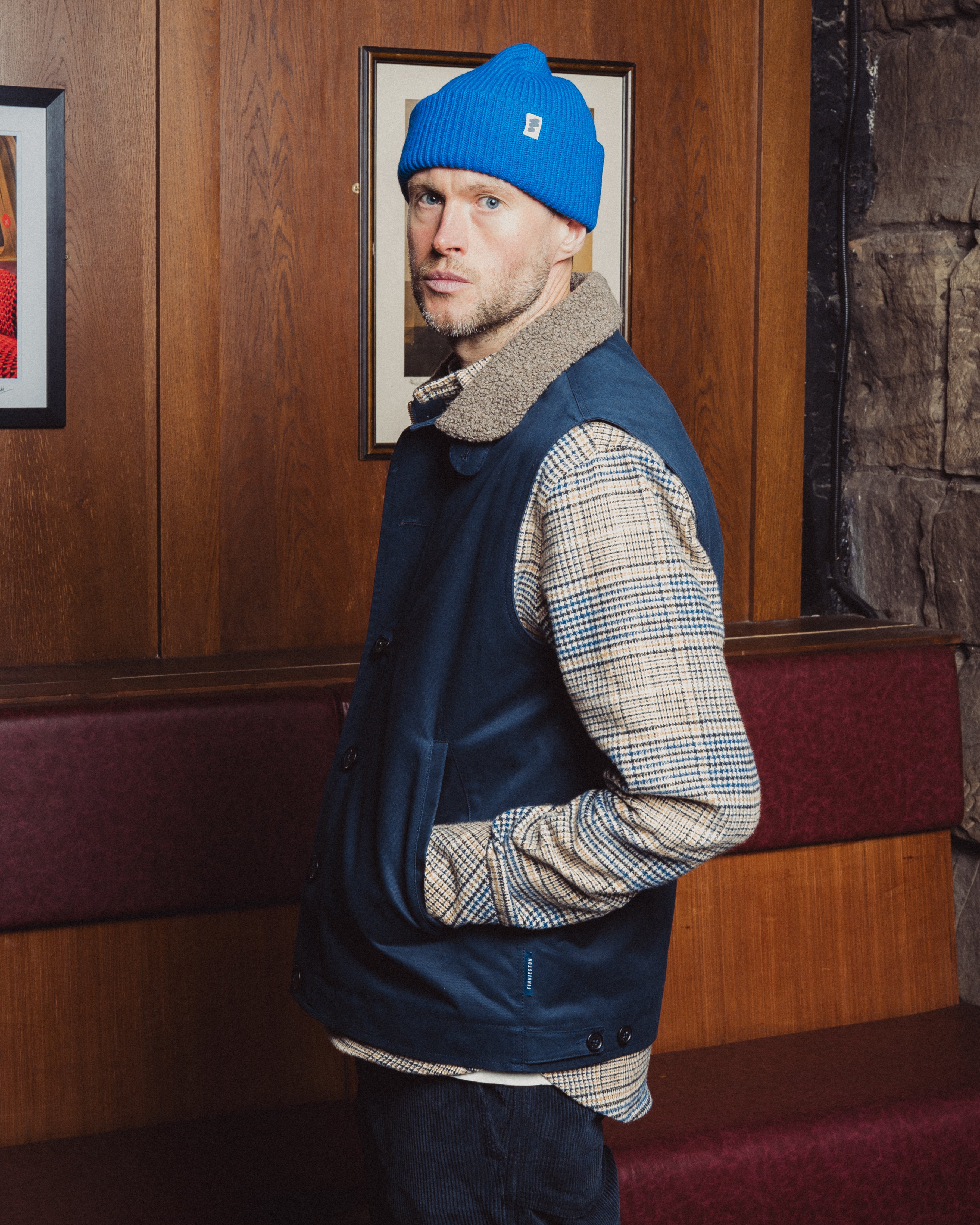 Man wearing a blue beanie and navy vest in a room with wooden paneling and framed pictures.
