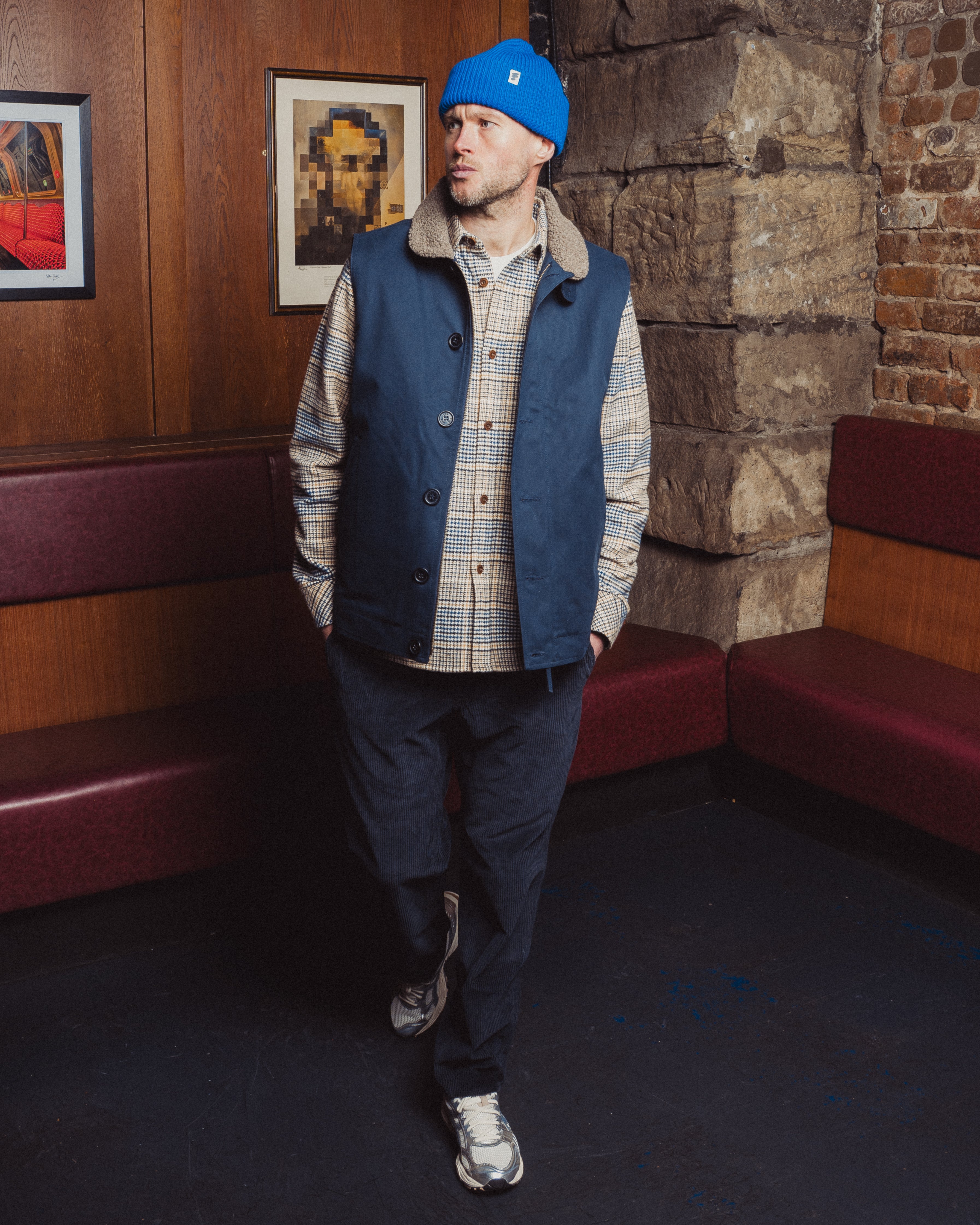 Man wearing a blue vest and plaid shirt standing in a room with brick walls and framed pictures.