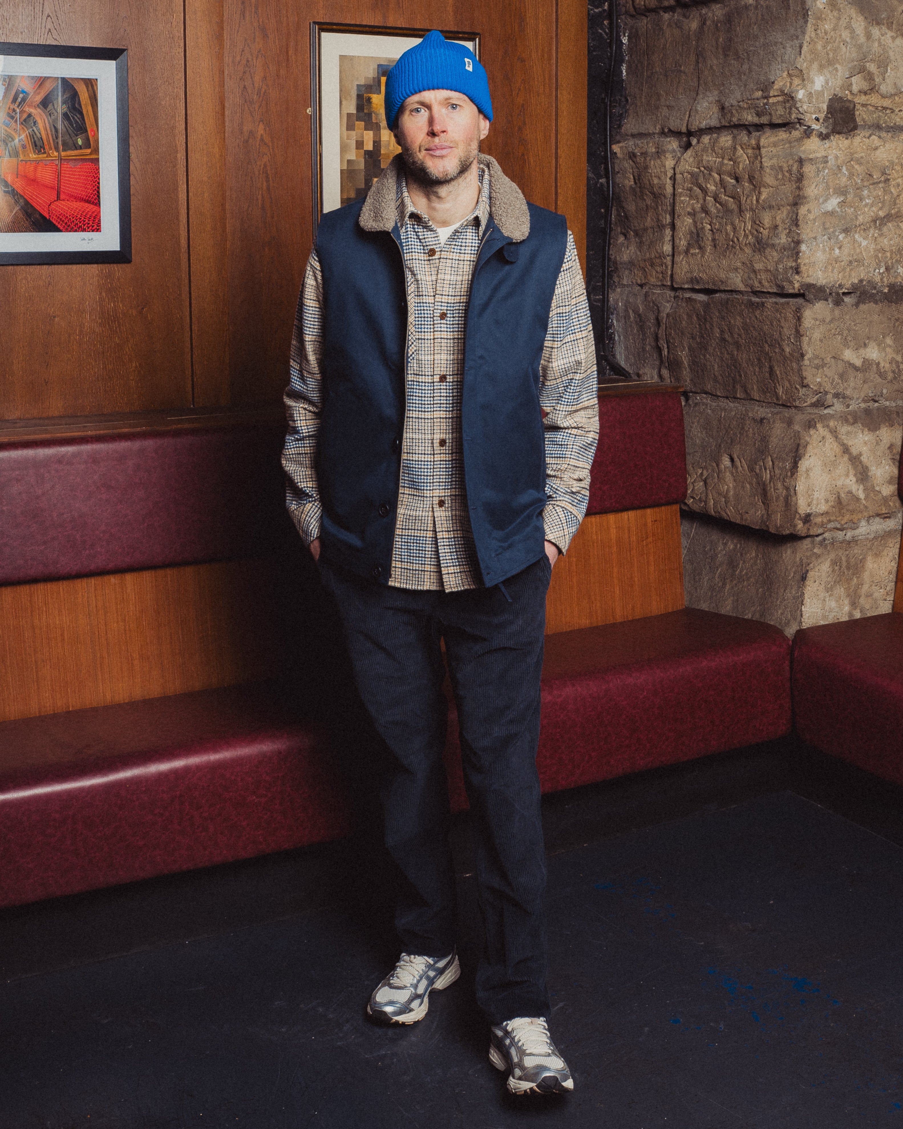 Man wearing a blue beanie and vest standing in a room with wooden walls and red benches.