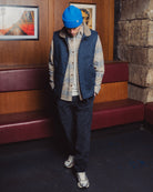 Person wearing a blue vest, plaid shirt, and dark pants standing in a room with wooden walls and a stone wall.