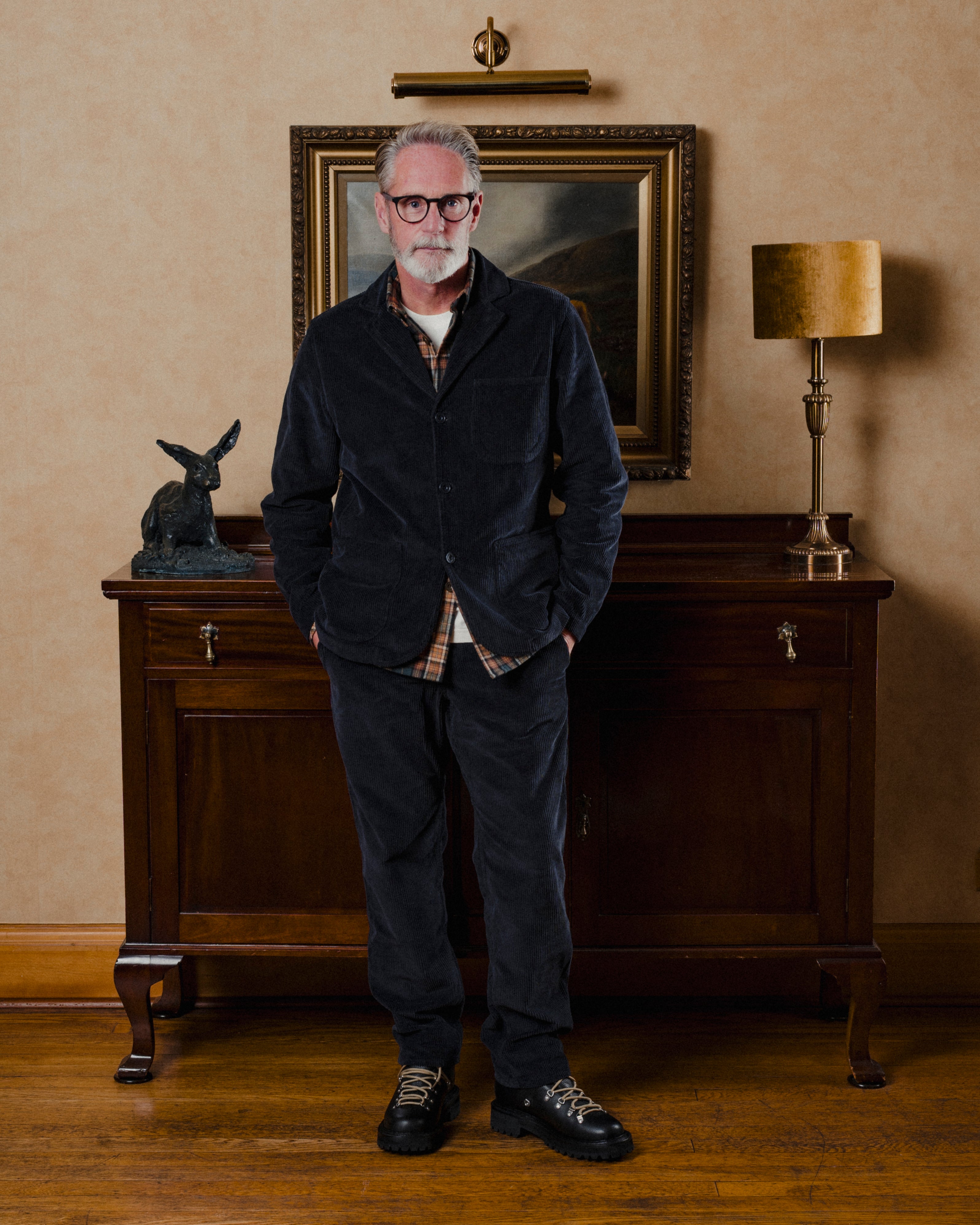 Man wearing navy corduroy suit standing in a room with a painting and lamp on a wall.